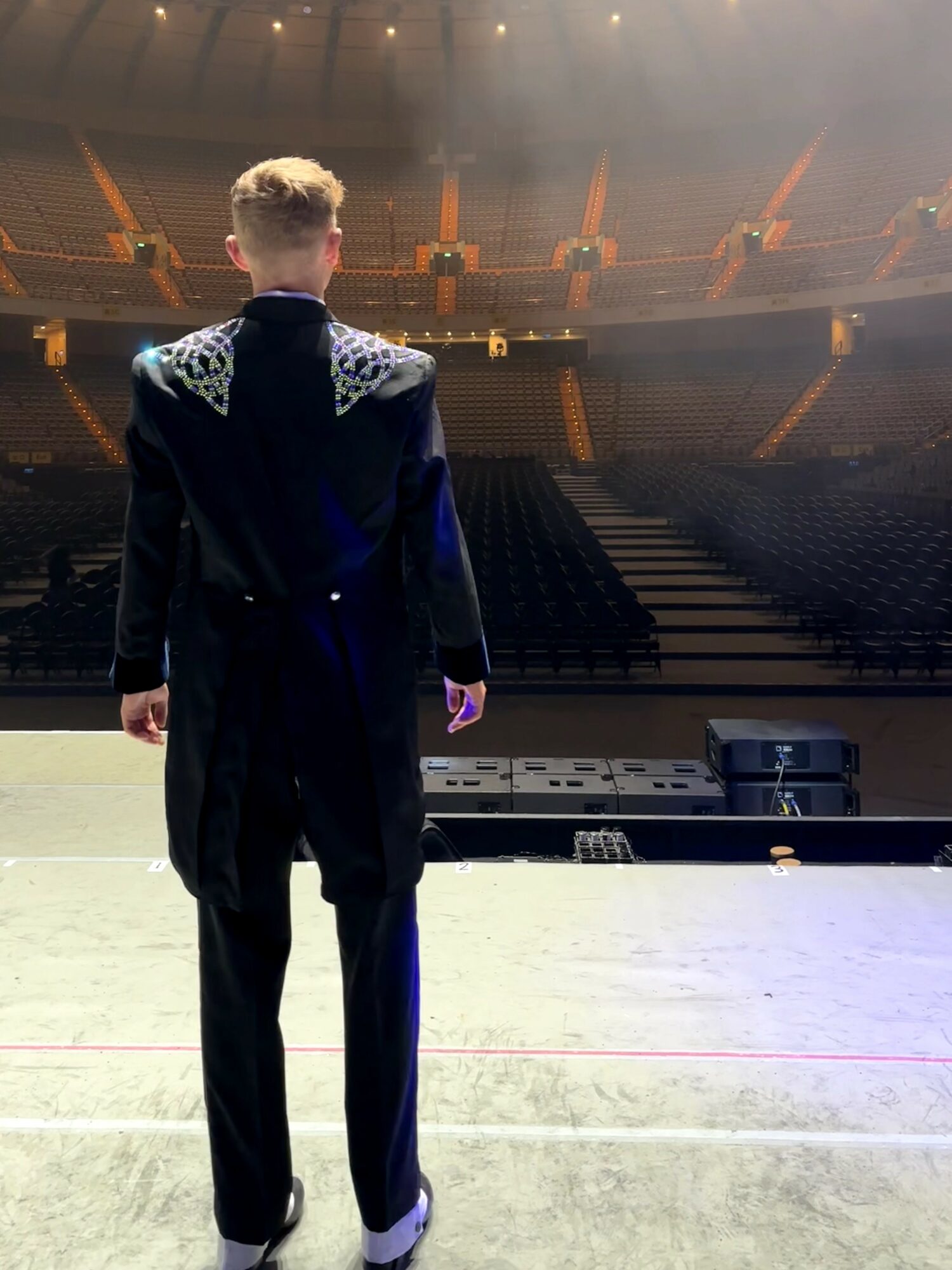Person standing on stage facing an empty auditorium with rows of seats, stage lights overhead, back view.