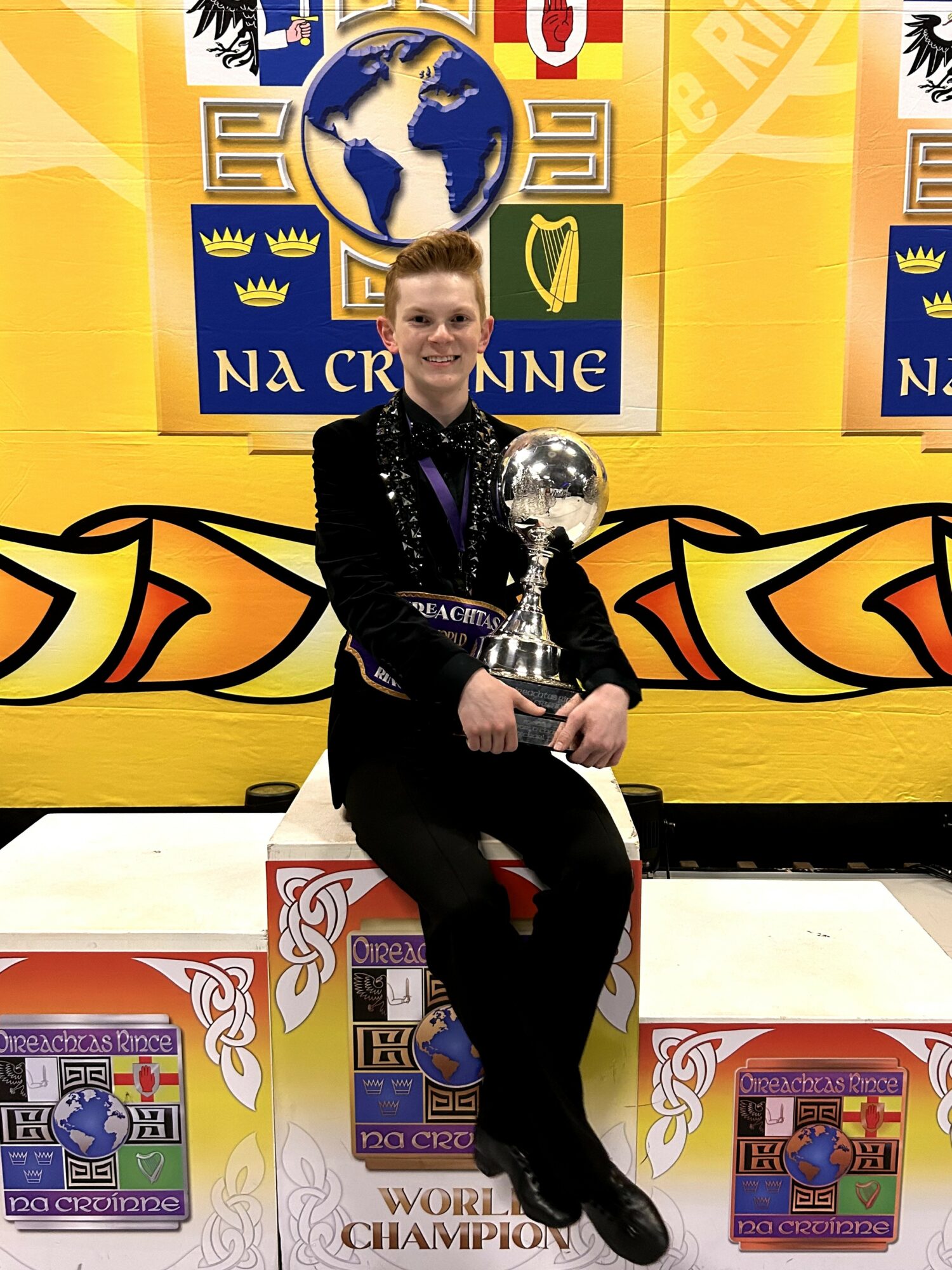 Person sitting on a podium holding a trophy, with a colorful backdrop featuring a globe and various symbols.
