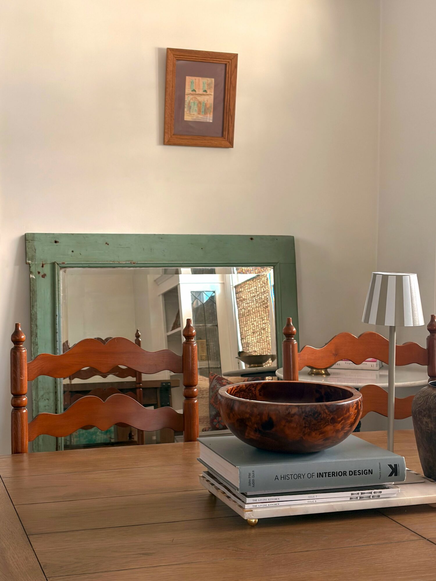 Dining table with chairs, a large mirror, a small lamp, and a stack of books in a room with a framed picture on the wall.