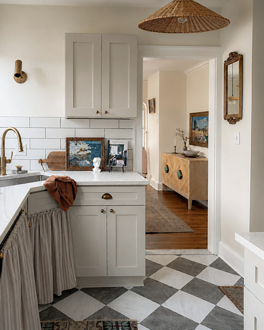 Kitchen corner with white cabinets, a gold faucet, and a wooden ceiling light, leading to a room with a wooden sideboard.