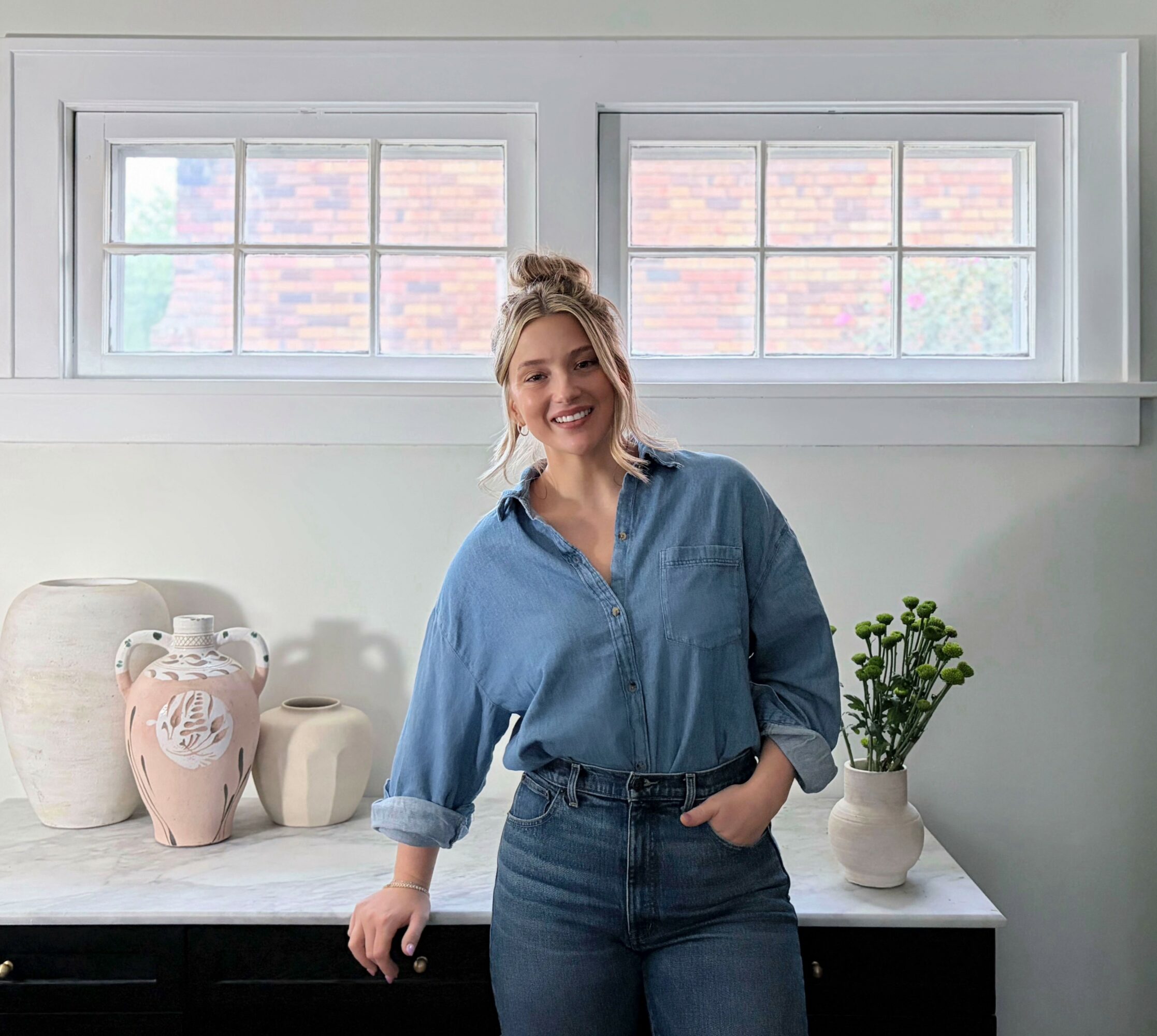 Smiling woman with blonde hair in a bun, wearing a blue shirt and jeans, standing in front of a window and a white countertop with vases and a plant.