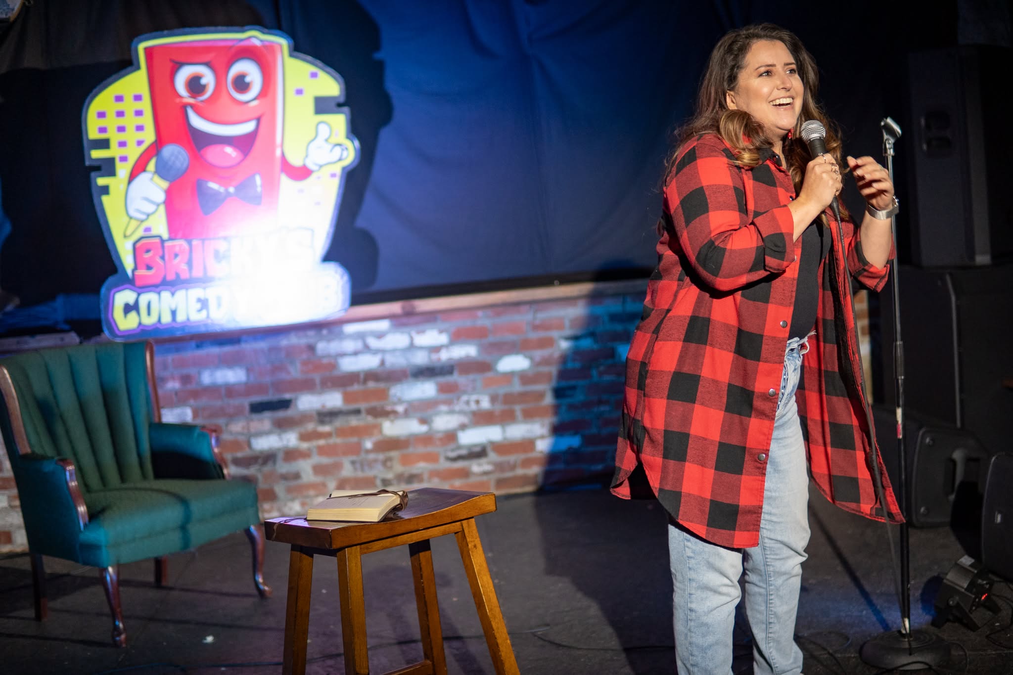 Woman performing stand-up comedy on stage with a brick wall background and a colorful cartoon logo on screen.