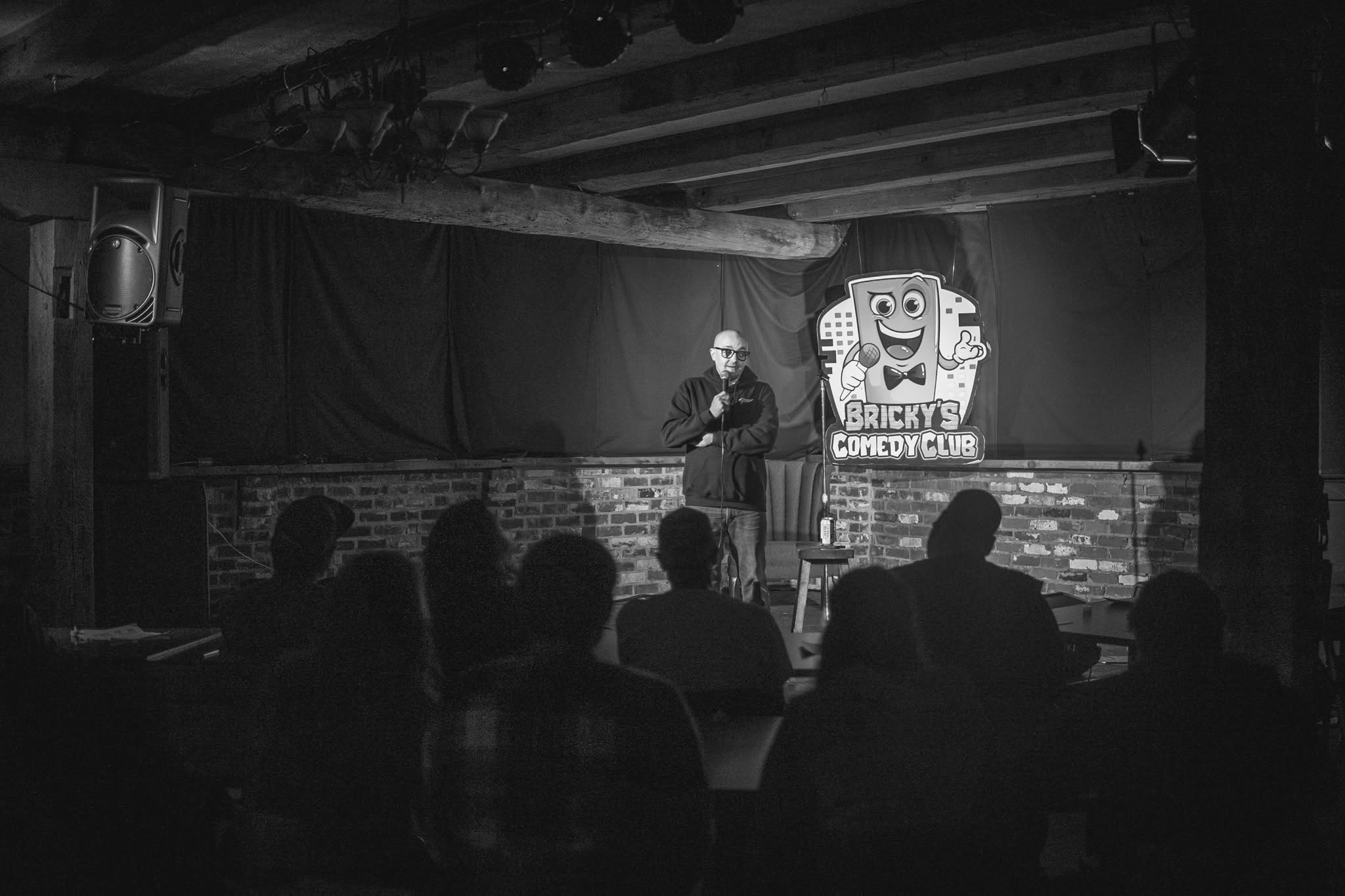 A person stands on stage with a microphone, performing in front of an audience in a comedy club setting.