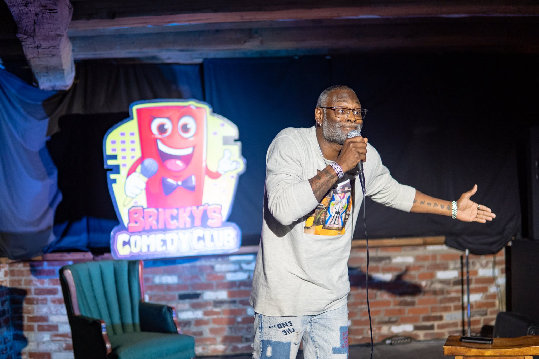 Man with glasses and beard speaking into microphone on stage with brick wall background, comedy club sign nearby.