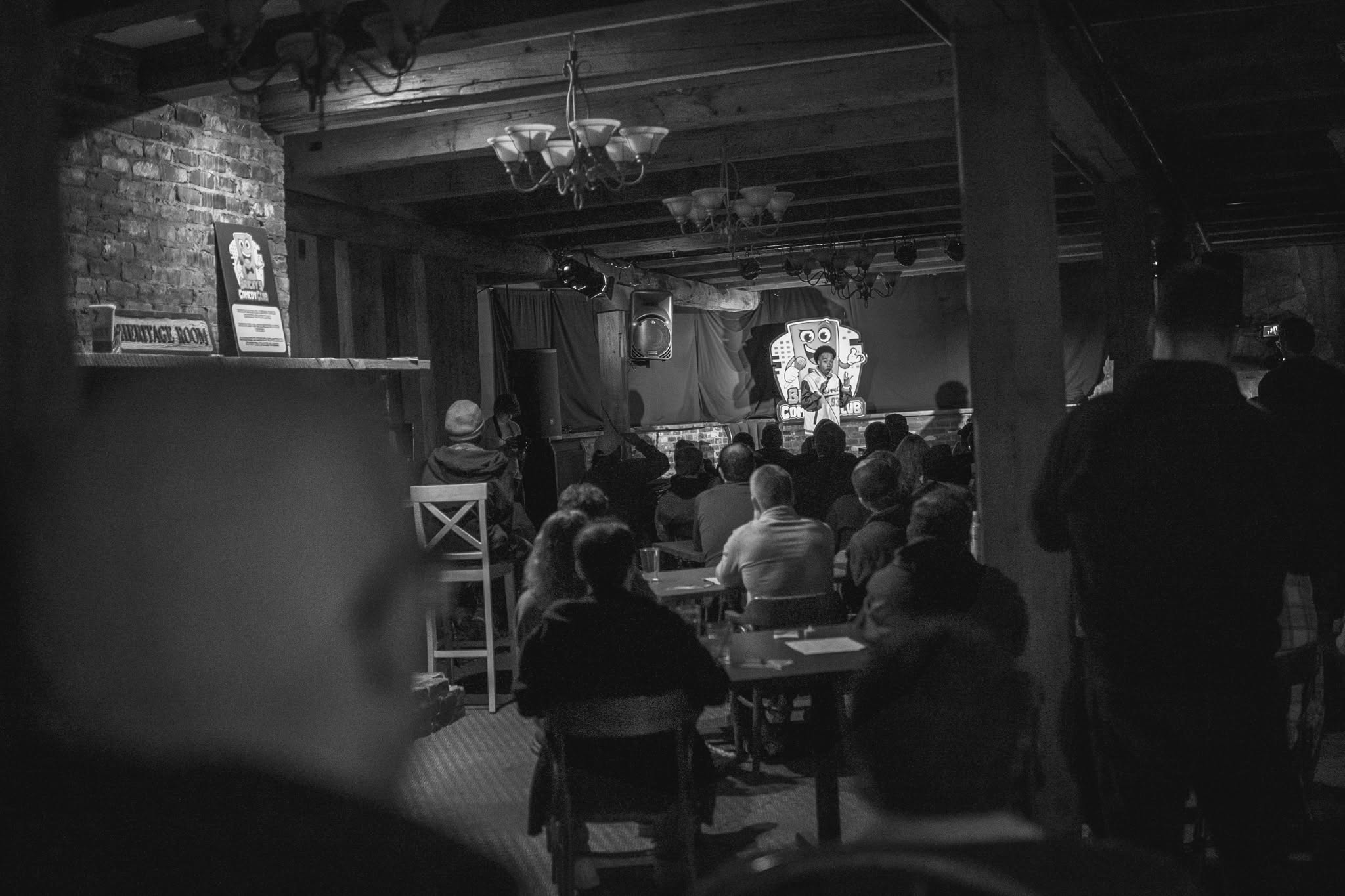 Audience watching a performance on stage in a dimly lit room with wooden beams and chandeliers.