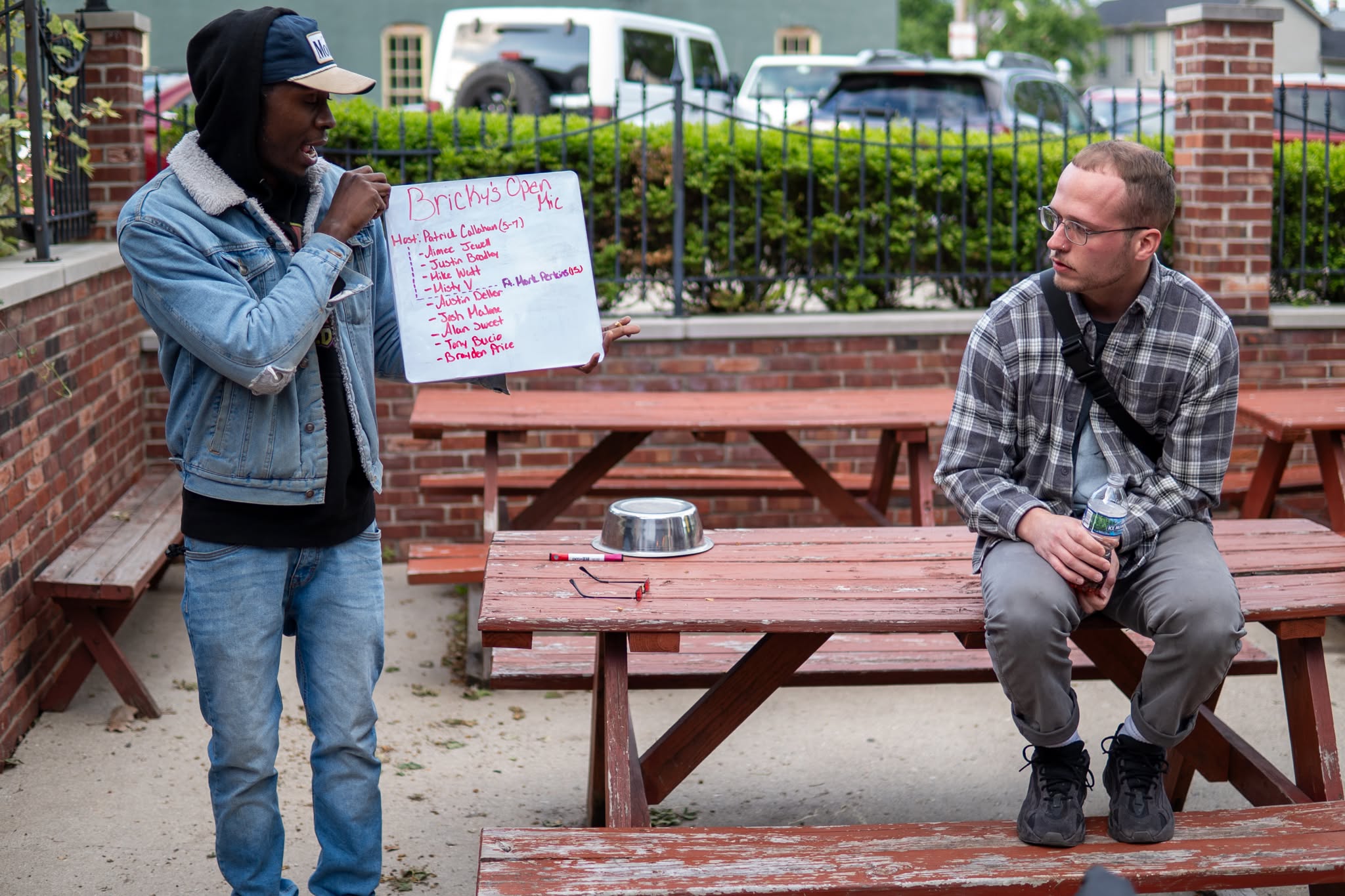 Two men outdoors near brick benches; one holds a whiteboard with writing, the other sits on a bench, wearing glasses and a plaid shirt.