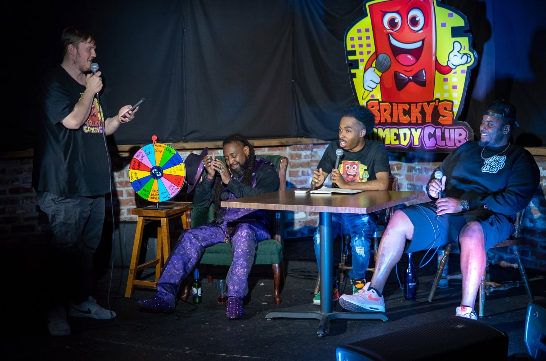 Person standing with microphone speaking to three seated people on stage at comedy club, colorful sign in background.