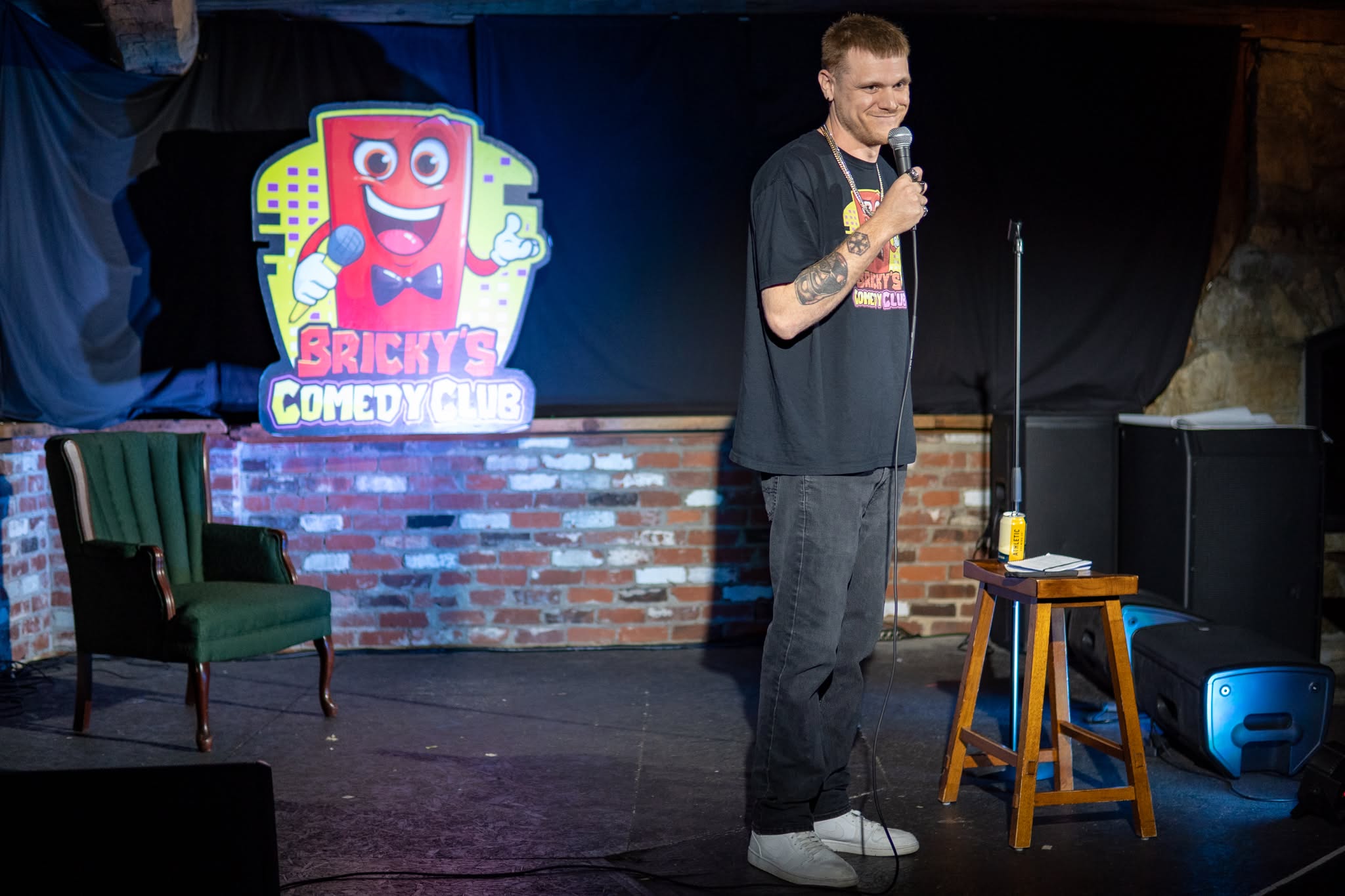 A man stands on stage holding a microphone, with a brick wall and a colorful Bricky Comedy Club sign behind him.