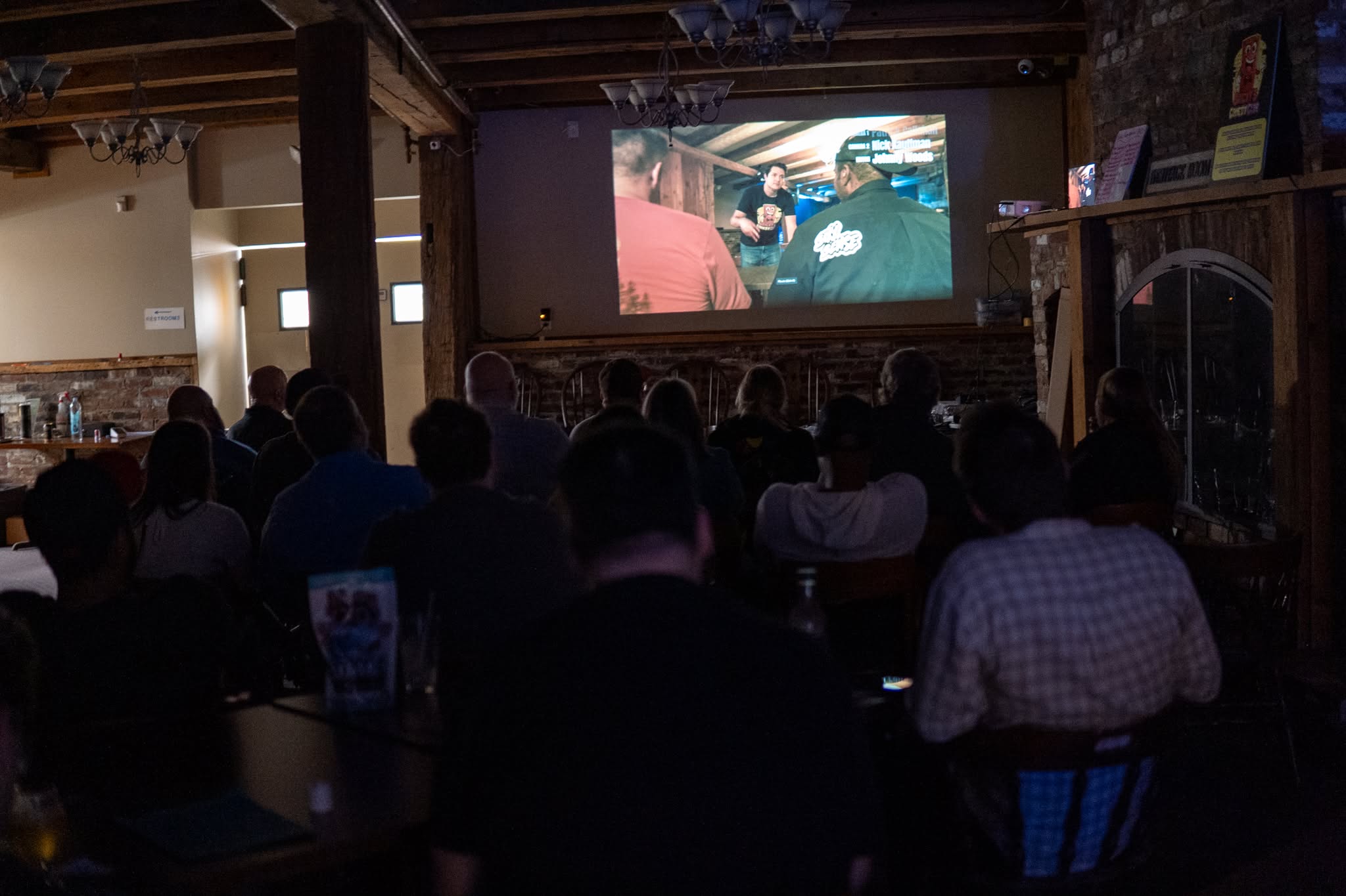 People watching a movie in a dimly lit room with a large screen, some seated at tables, others standing, wooden beams overhead.