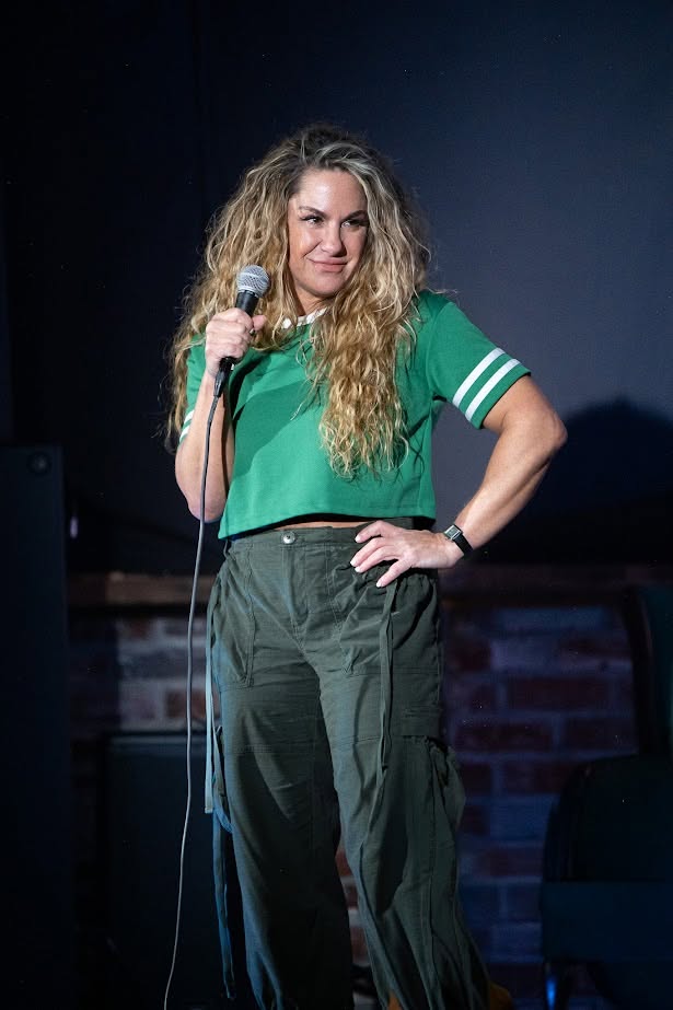 Woman with long curly hair holding a microphone on stage, wearing a green shirt and dark pants, smiling confidently.