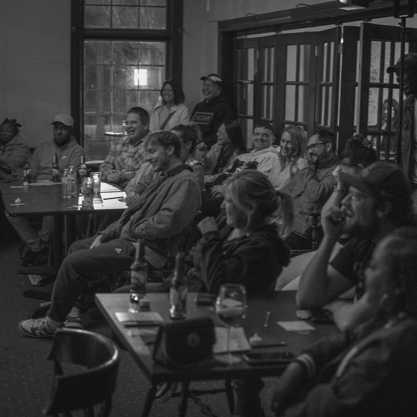 Group of people seated at tables in a room, some smiling and listening, with large windows in the background.