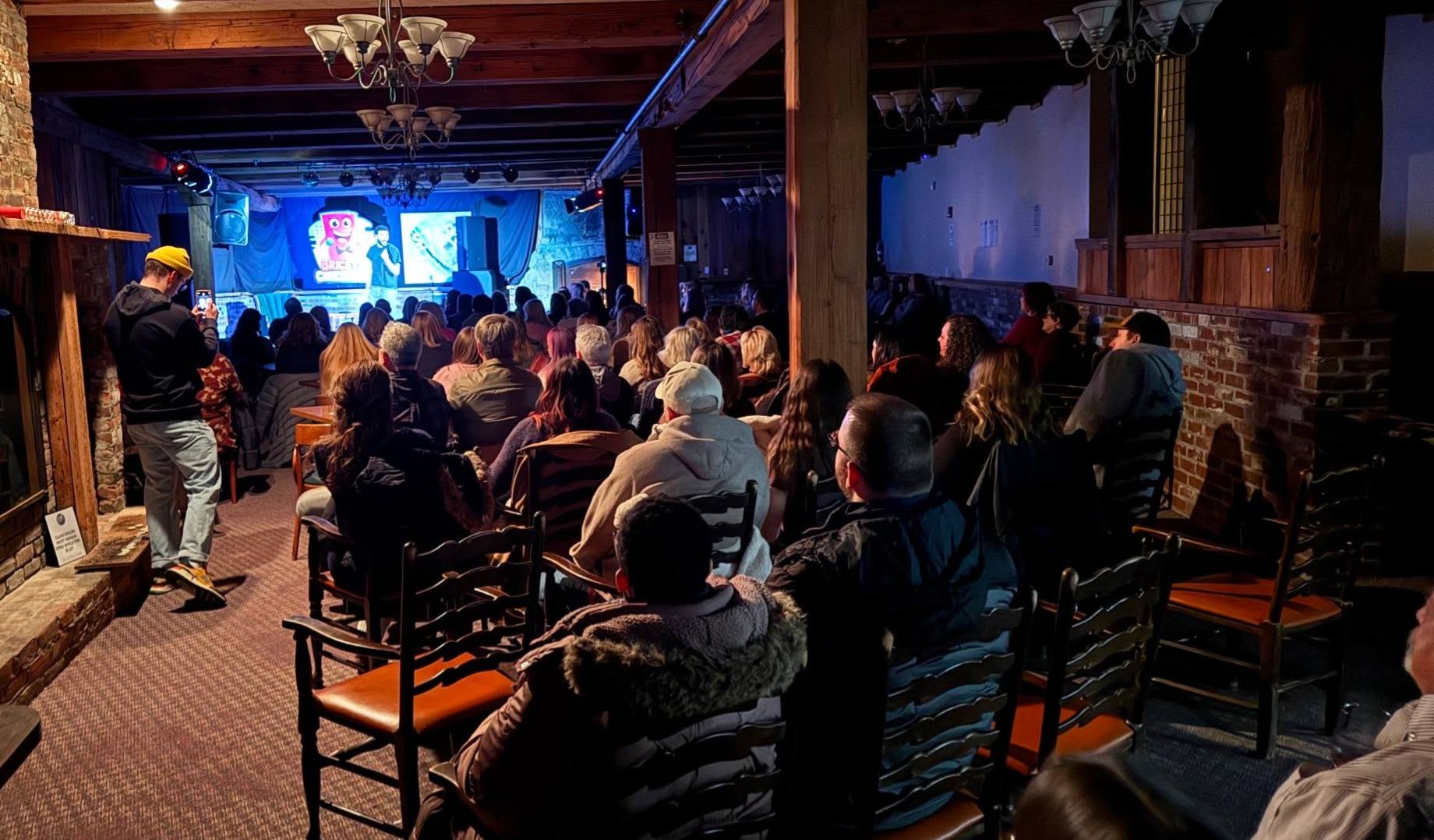 Audience watching a performance on stage in a dimly lit venue with wooden beams and brick walls.