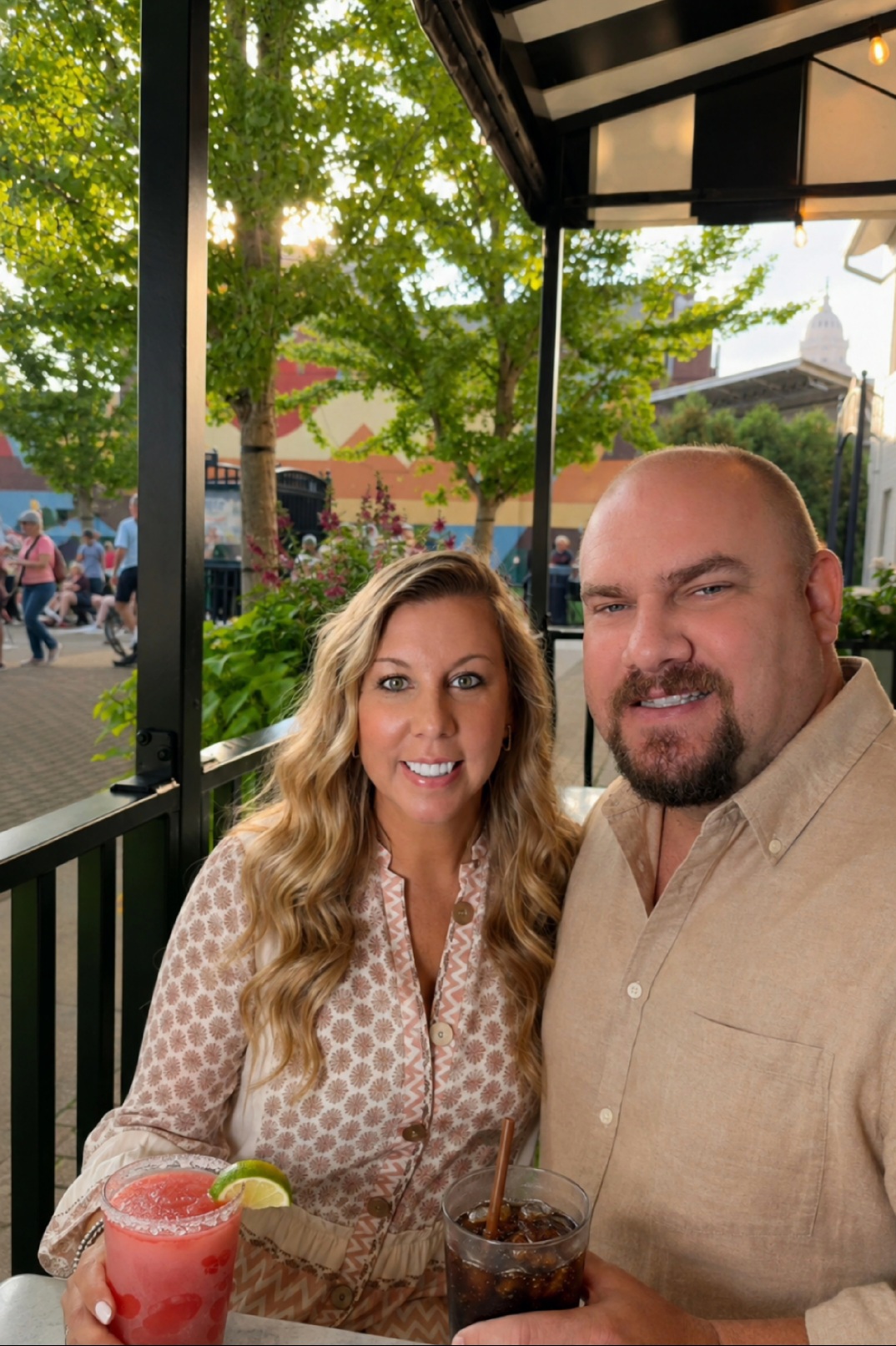 Two people sitting at a table outdoors, smiling, with drinks, trees, and a crowd in the background.