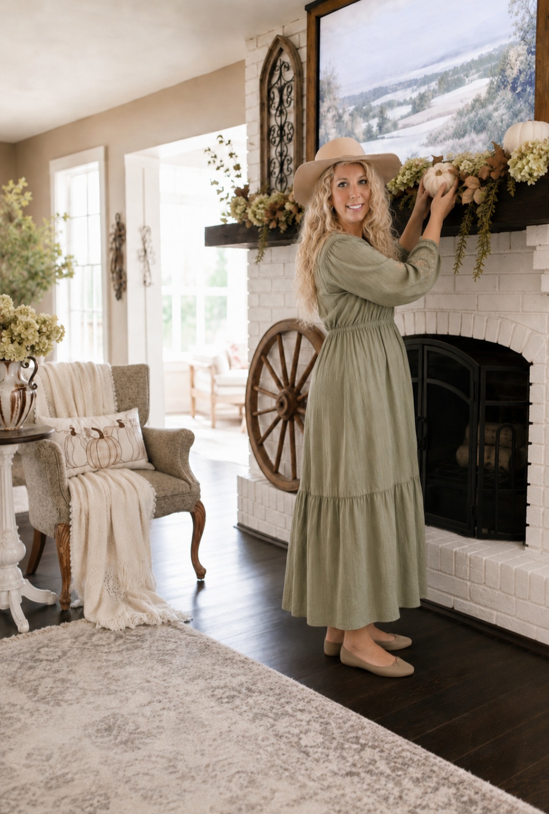 Woman in a long dress and hat decorates a fireplace mantel with flowers in a cozy living room.