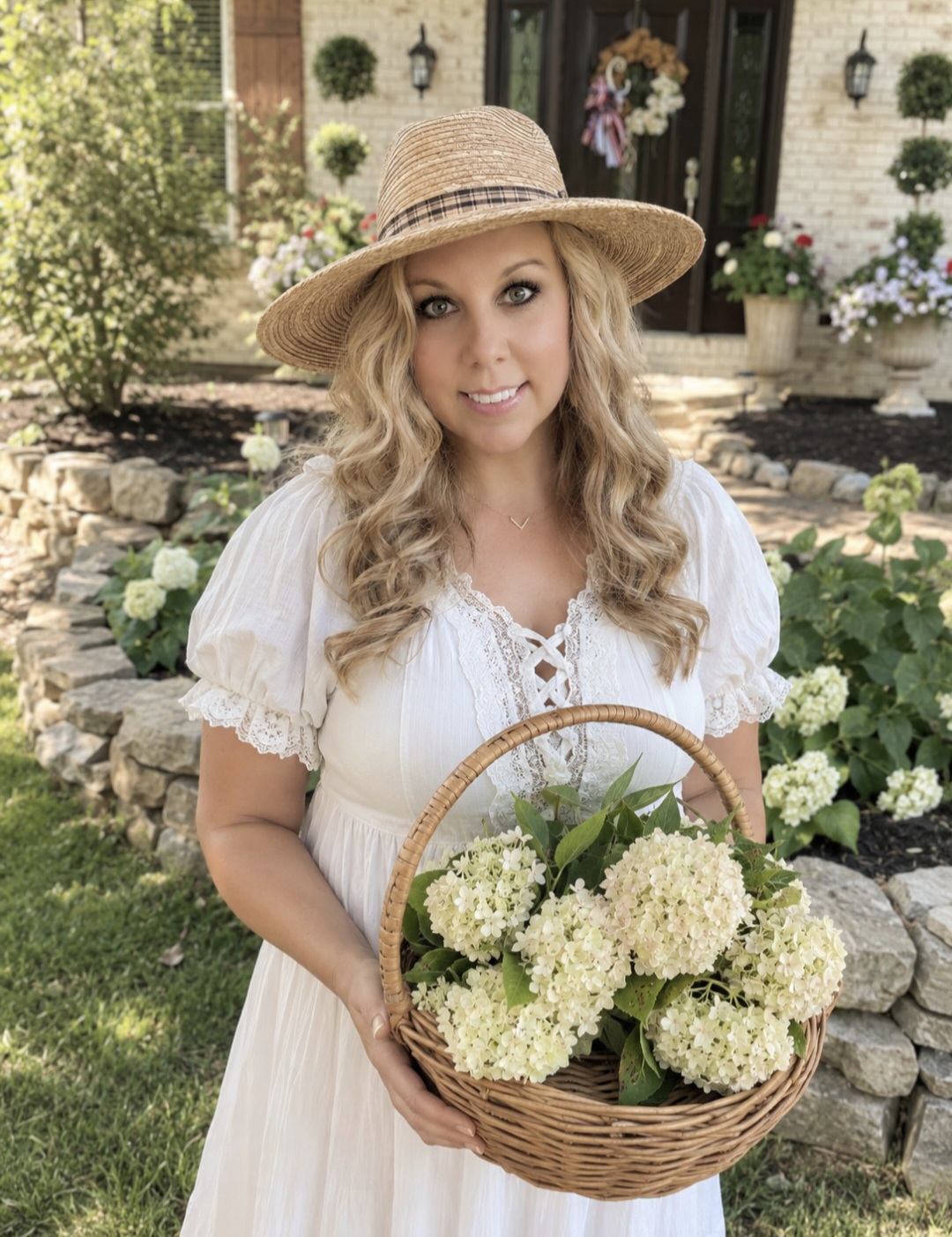 Woman in a white dress and wide-brimmed hat holding a basket of white hydrangeas outdoors.