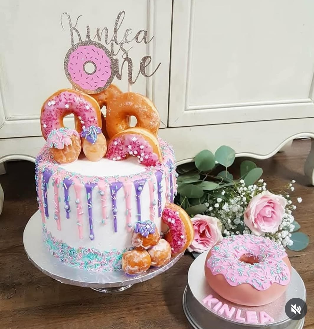Cake decorated with donuts, sprinkles, and a pink donut topper, with flowers and a sign in the background.