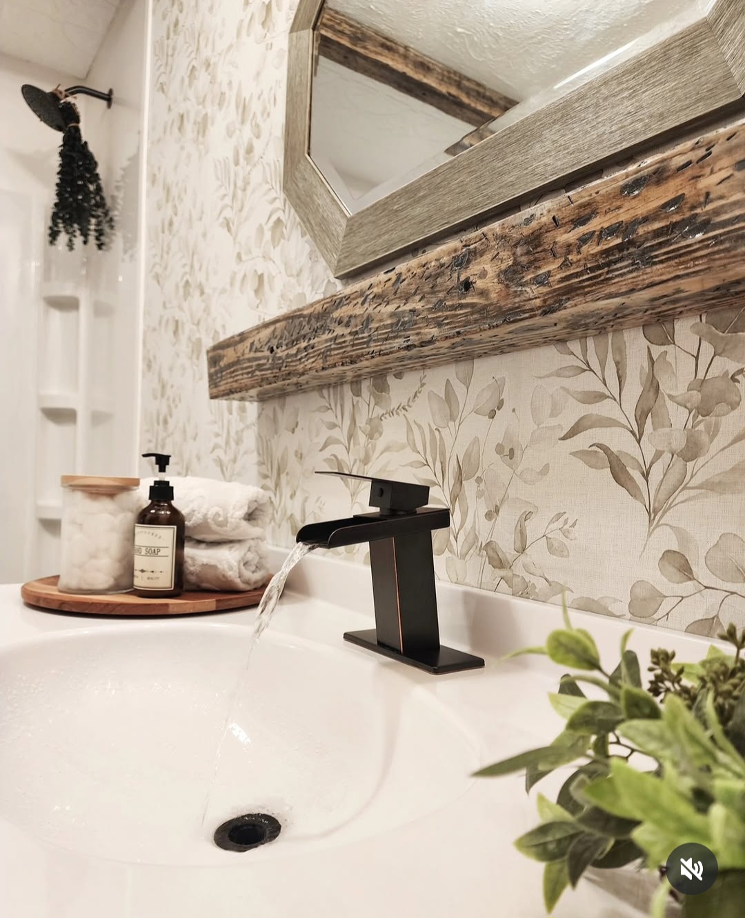 Bathroom sink with faucet, mirror, plant, and toiletries on a wooden tray, wall with floral wallpaper, and towel.