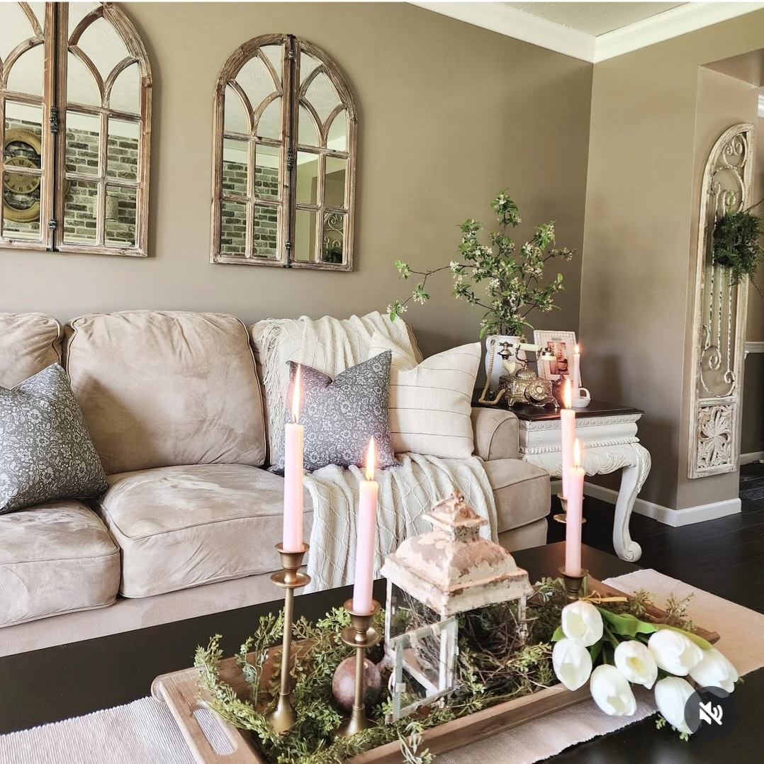 Living room with beige sofa, decorative pillows, candles, and a tray with flowers and greenery, wall mirrors, and a plant in the corner.