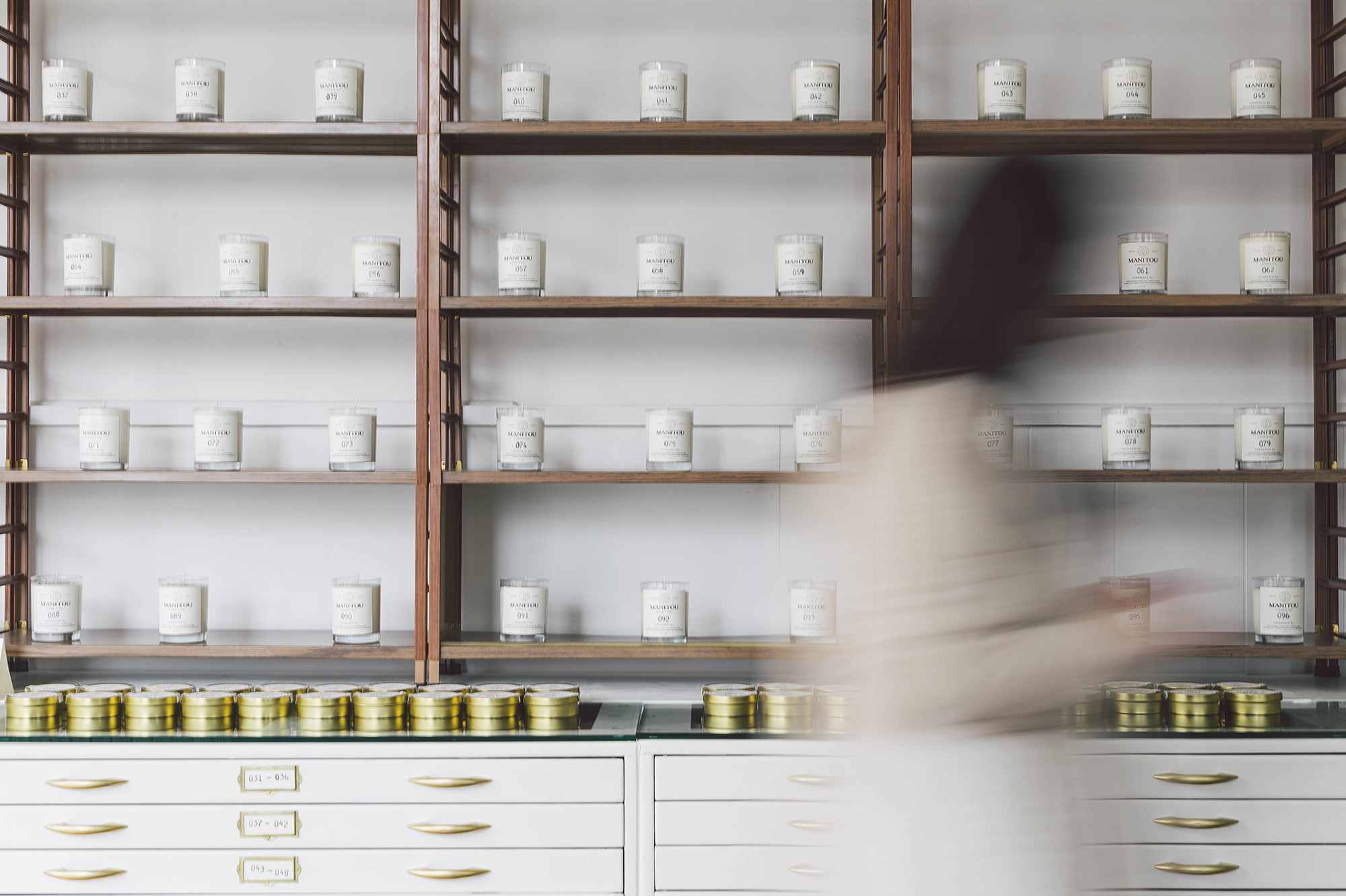 Blurred person walking past shelves with jars and containers in a store or pharmacy.