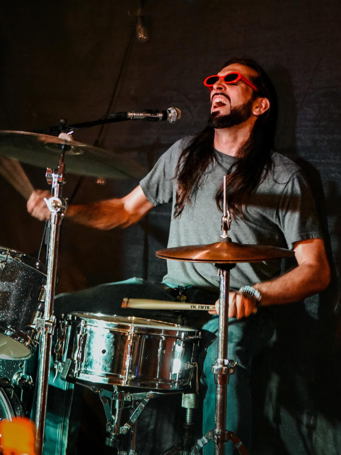 Musician playing drums with long hair, beard, sunglasses, and a gray shirt, smiling and singing into a microphone.