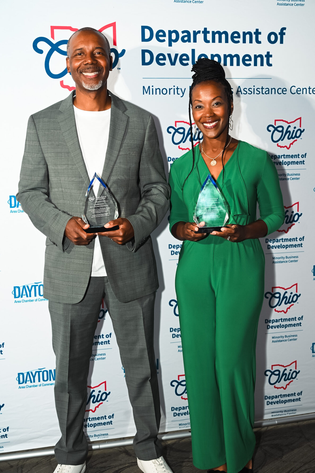 Two people holding awards, standing in front of a backdrop with logos and text, smiling. One man in a gray suit, one woman in a green outfit.