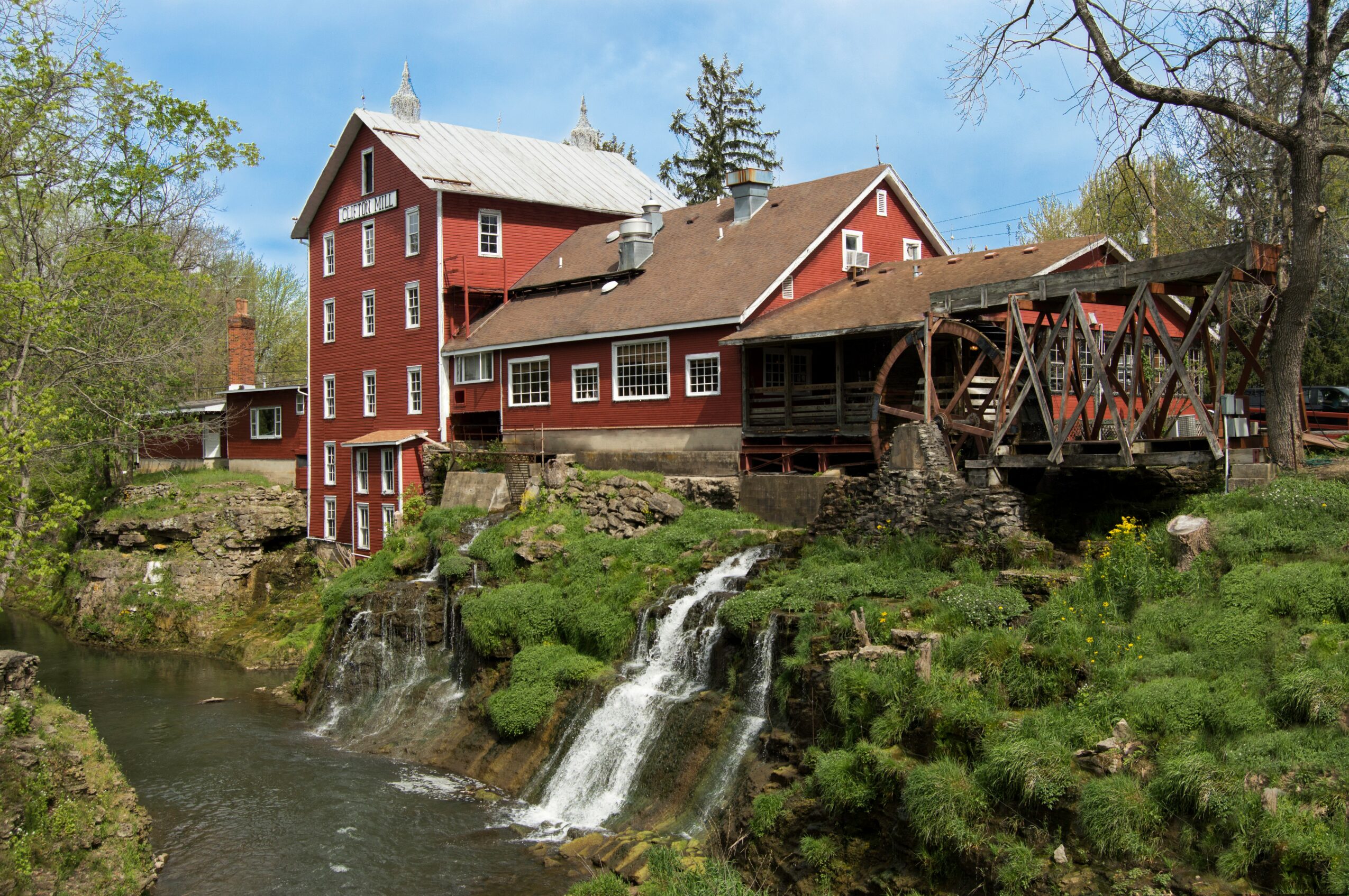 Red mill building with water wheel beside a small waterfall and river, surrounded by trees and greenery.