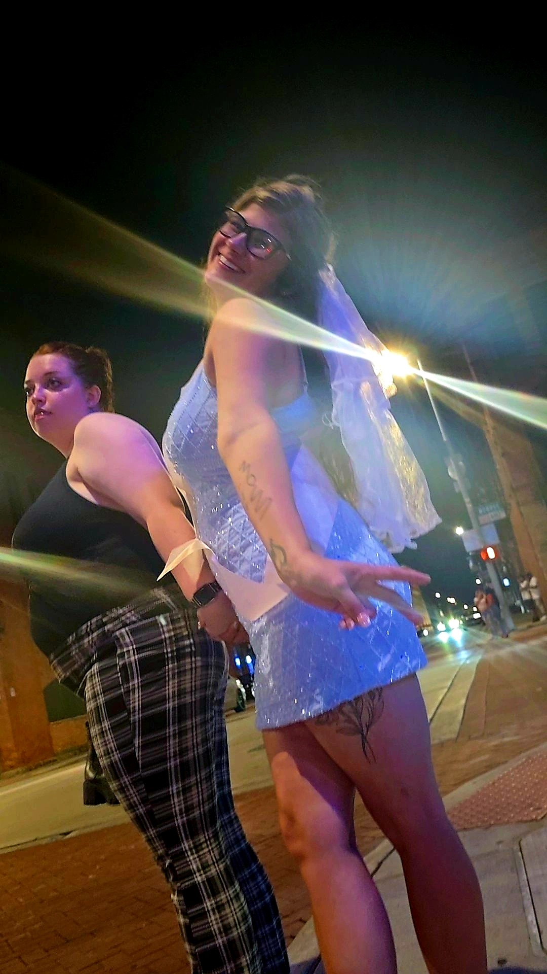 Two women standing outdoors at night, one in a dress and the other in plaid pants, with city lights in the background.