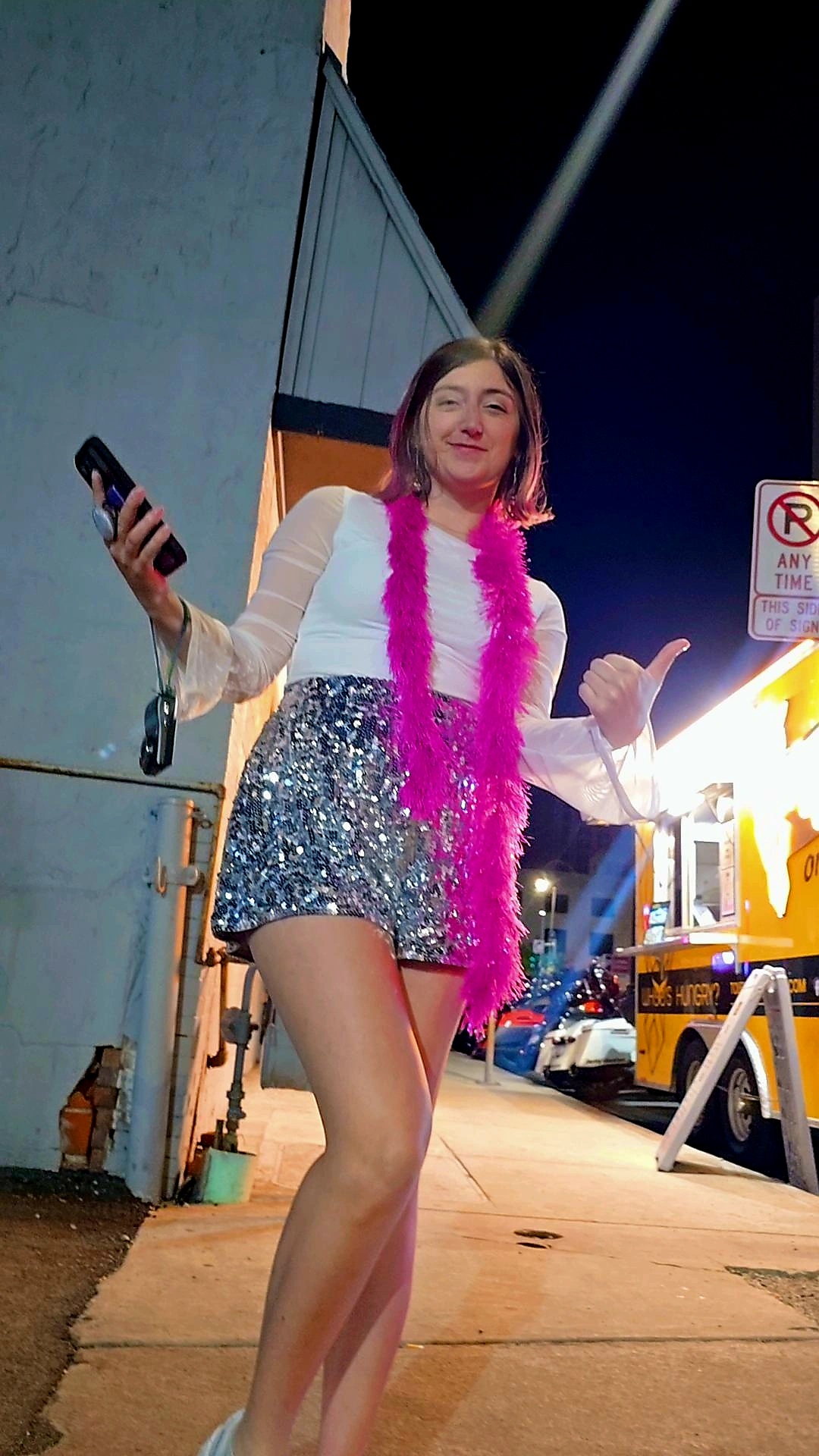 Young woman standing outdoors at night, wearing a white top, shiny shorts, pink feather boa, holding a phone, smiling, with streetlights and vehicles in background.