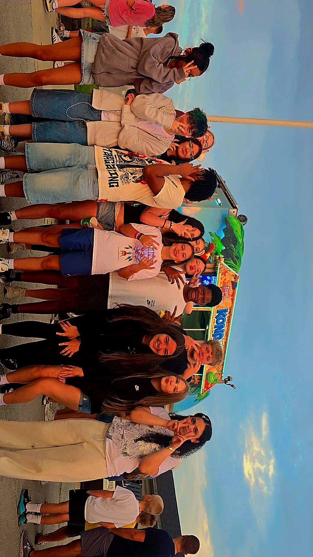 Group of people standing outdoors with a blue sky and clouds, some holding umbrellas, in casual clothing.