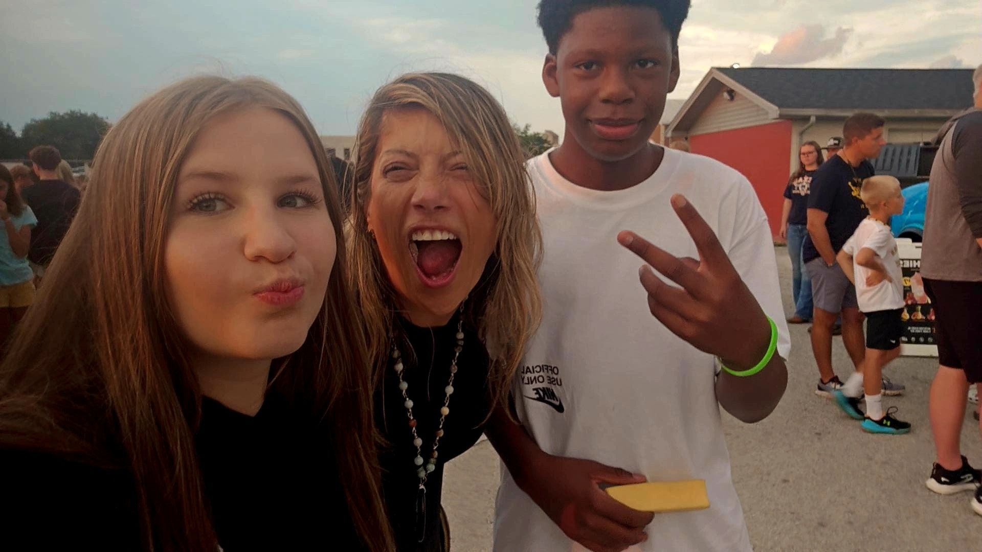 Two young women taking a selfie outdoors, one making a peace sign, the other pouting, with people and buildings in background.