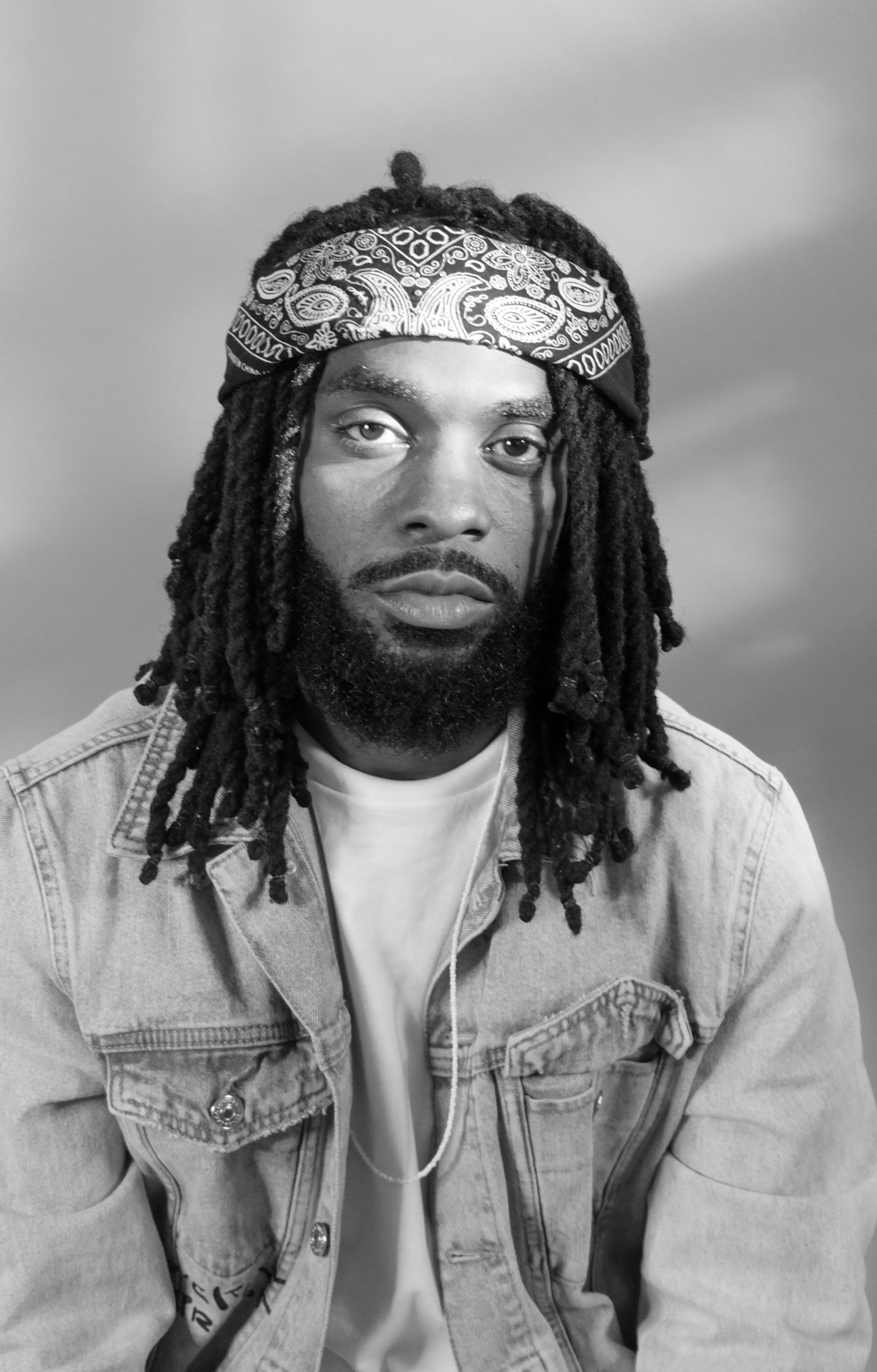 Black and white portrait of a man with dreadlocks wearing a bandana and denim jacket.