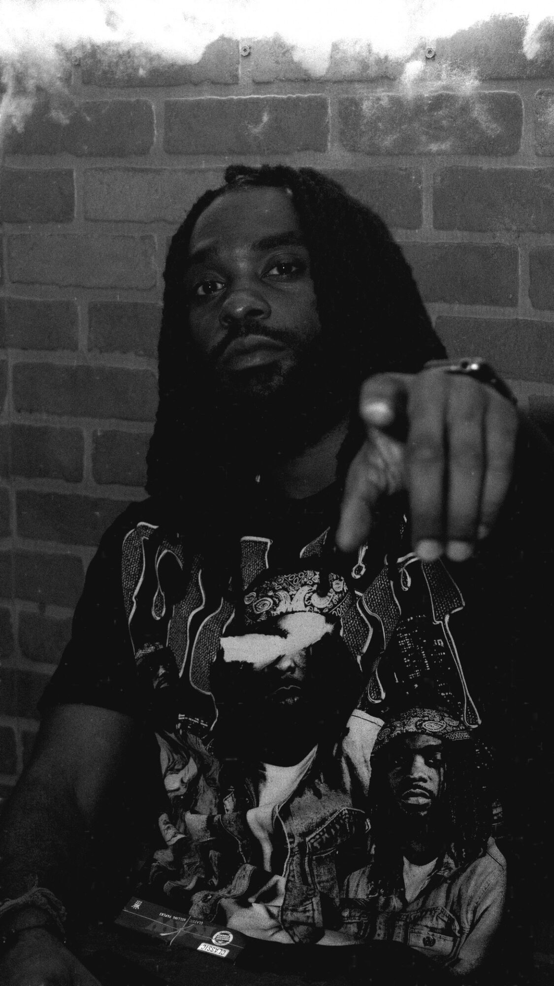 Person with long dreadlocks pointing at the camera, standing against a brick wall, in black and white.