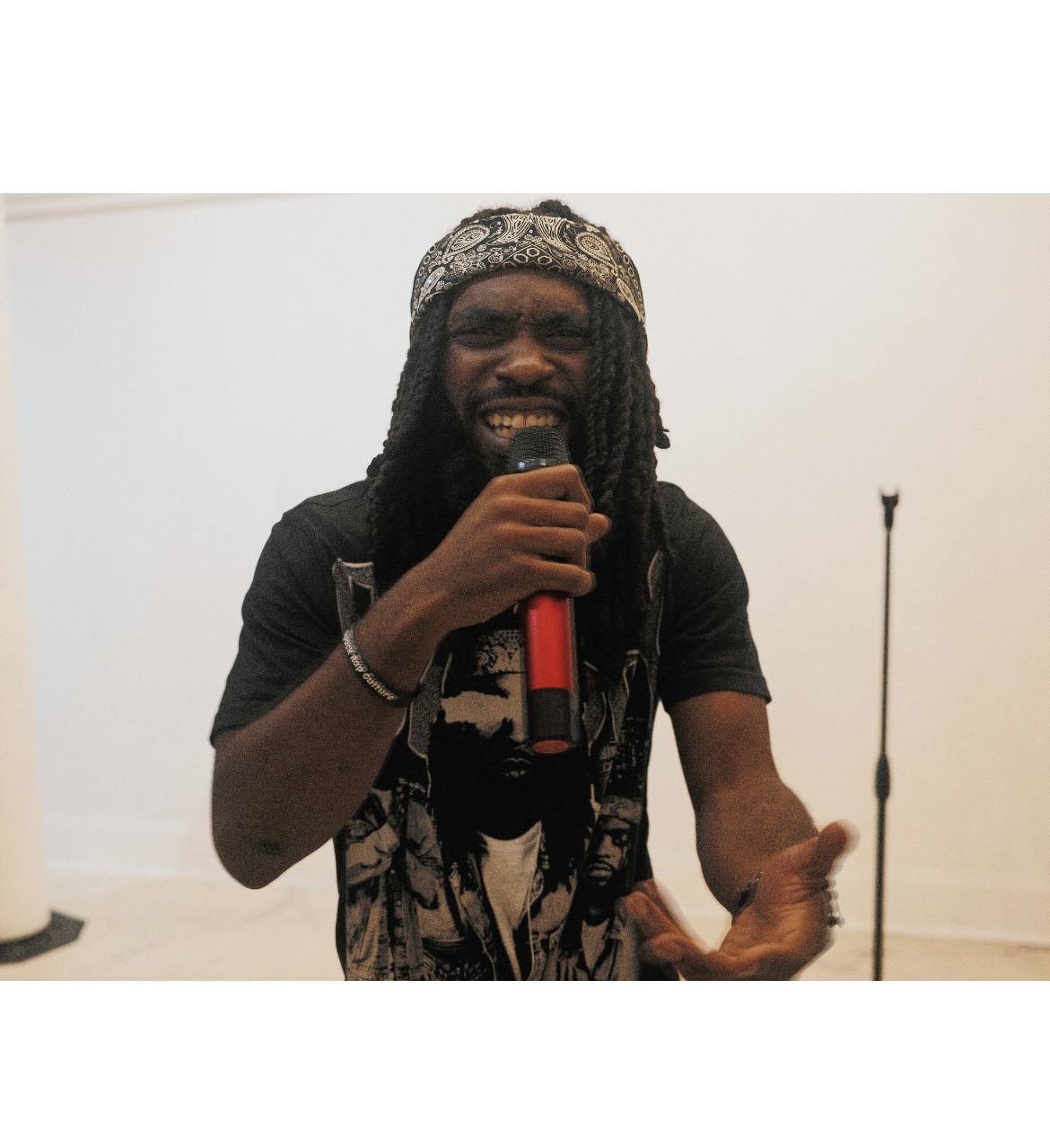 Person with dreadlocks and headband holding a microphone, smiling, in a room with a white wall.