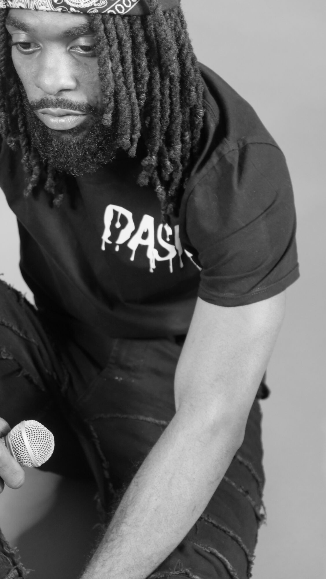 Man with dreadlocks and beard wearing a black T-shirt, sitting with arm extended, holding a microphone, in black and white.
