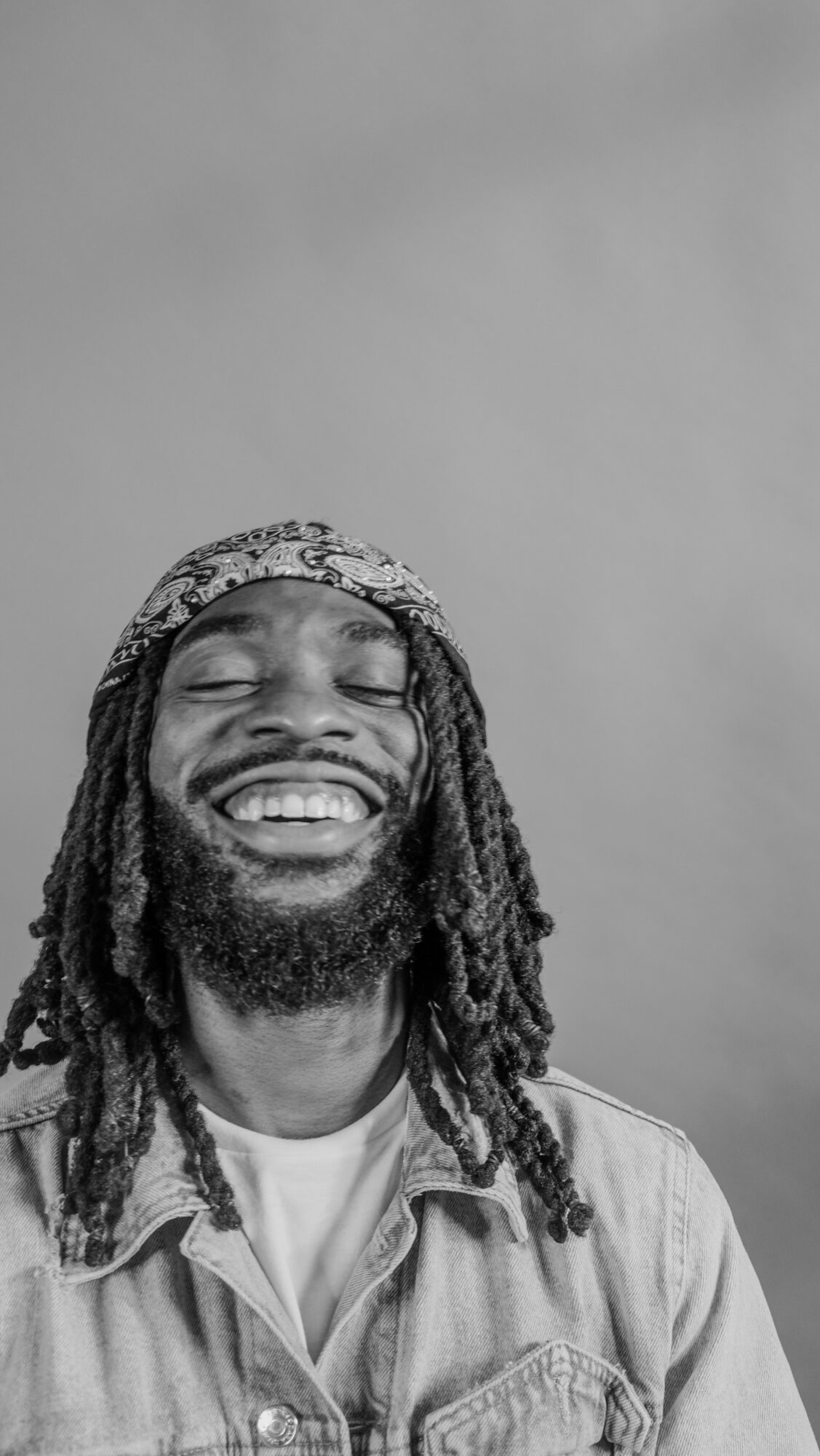 Person with dreadlocks and beard smiling, wearing a denim jacket, against a plain background.