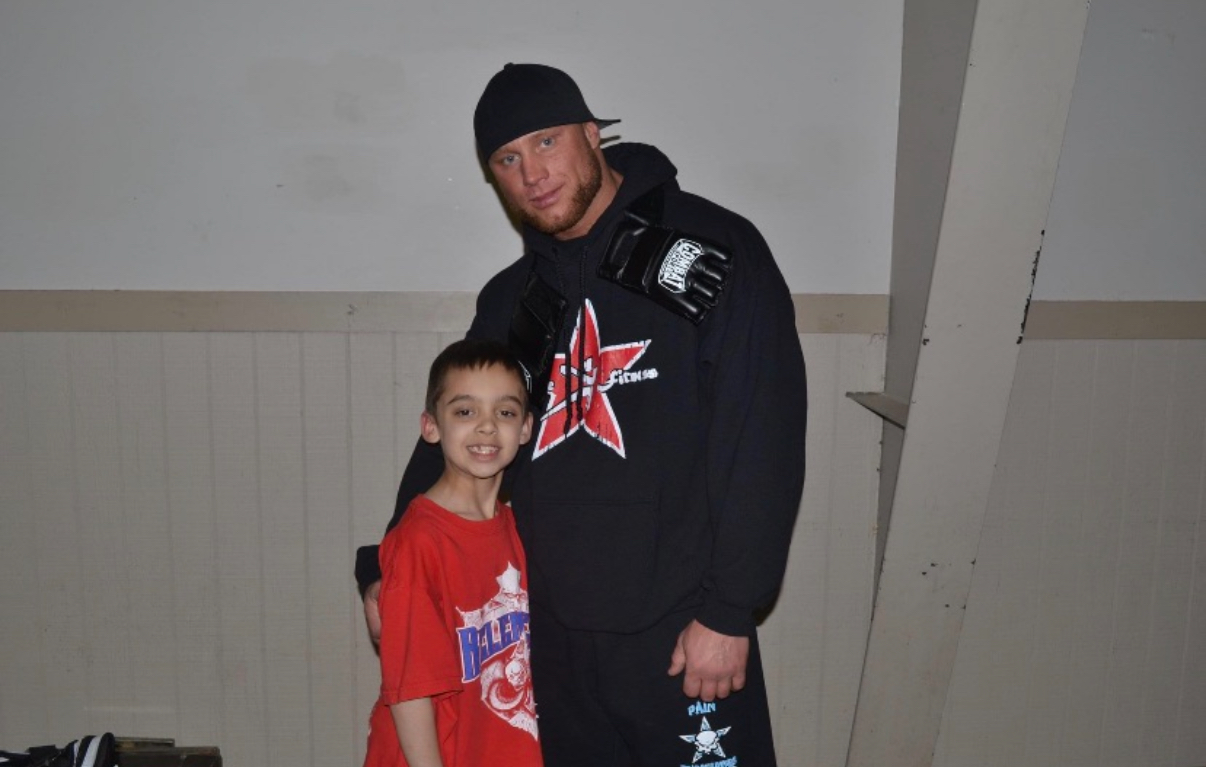 A man and a child stand together indoors, smiling at the camera, with a plain wall background.