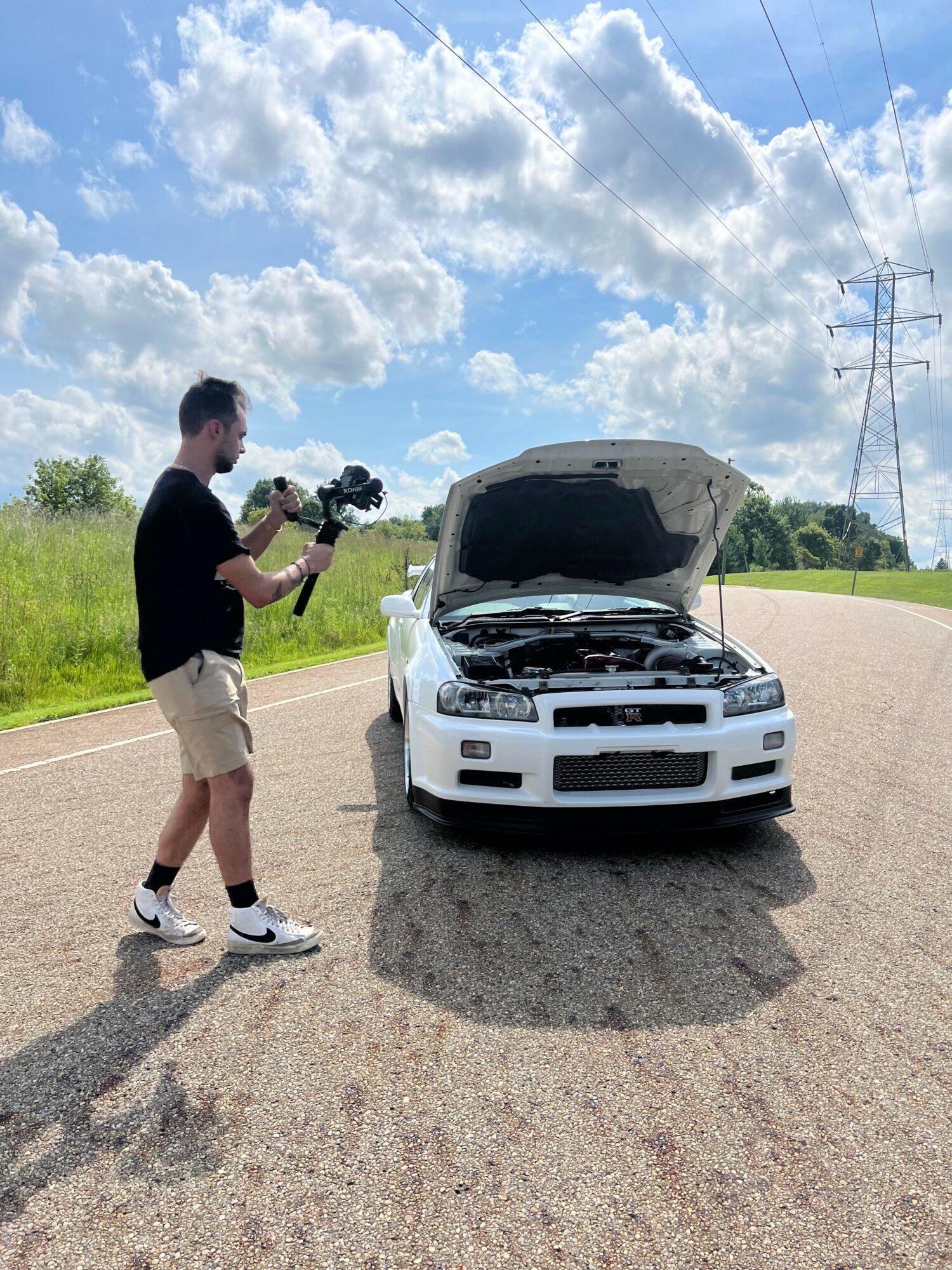Person standing on a road taking a photo of a white car with a person inside, under a partly cloudy sky.