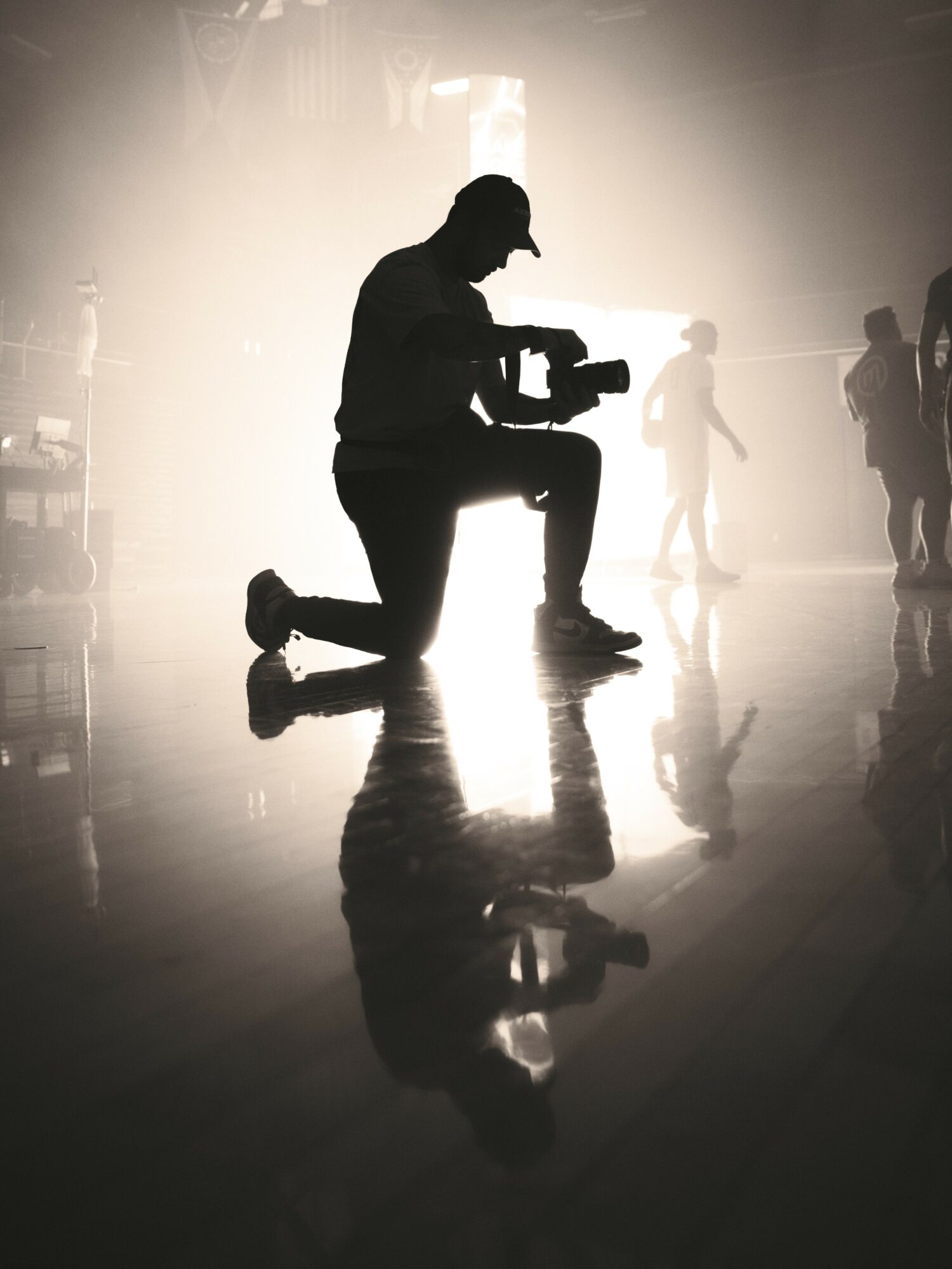 Person kneeling with a camera, silhouette against bright background, reflections on the floor, other people in the distance.