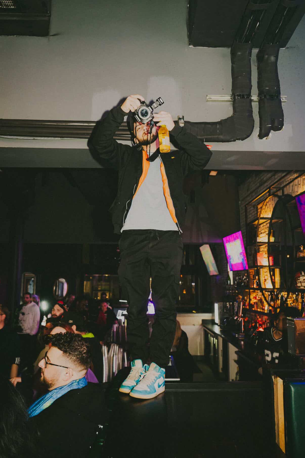 Person standing on a bar counter taking a photo with a camera, in a dimly lit bar or club setting.