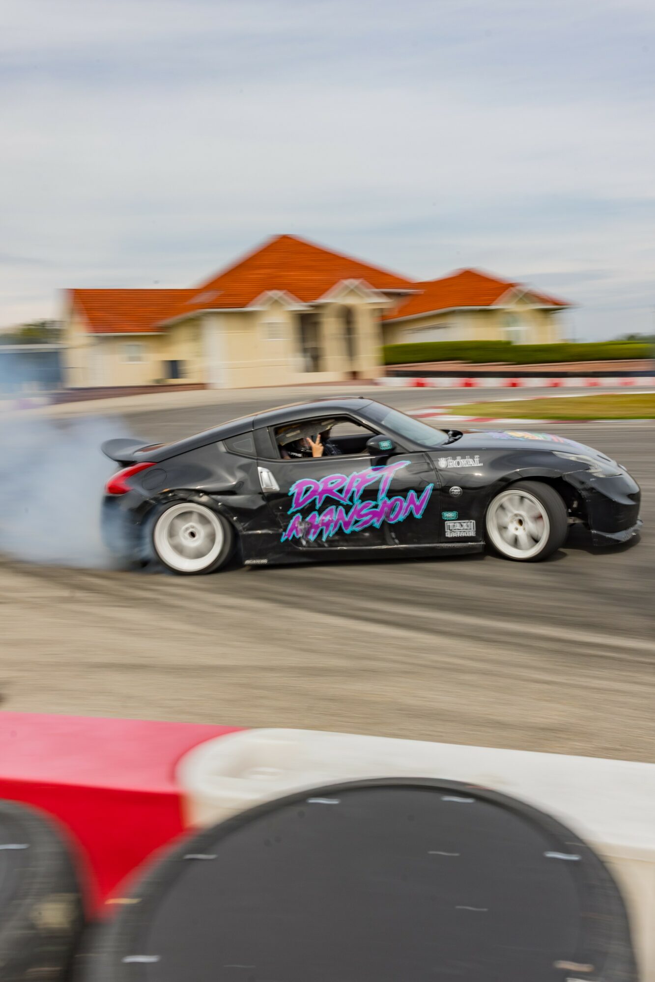 Black sports car with purple and pink graphics speeding on a track, blurred background of buildings and sky.