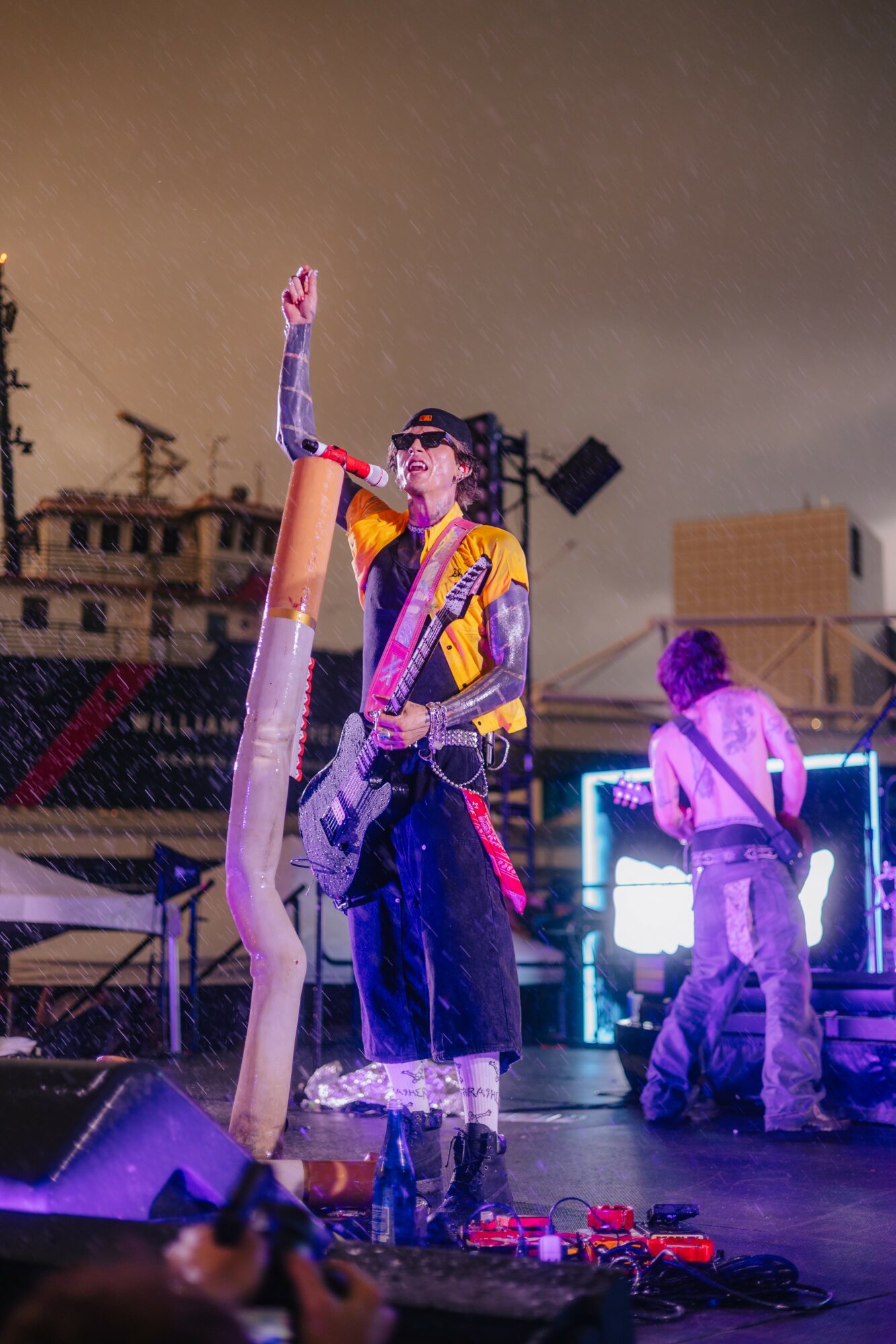 Performer on stage with a guitar, raising one hand, with another person in the background, stage lights, and equipment.