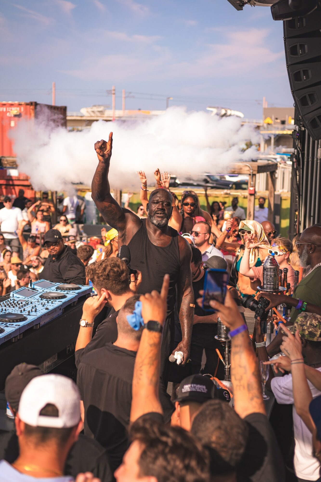 Crowd at outdoor event with a person raising a hand, smoke in the background, and people taking photos.