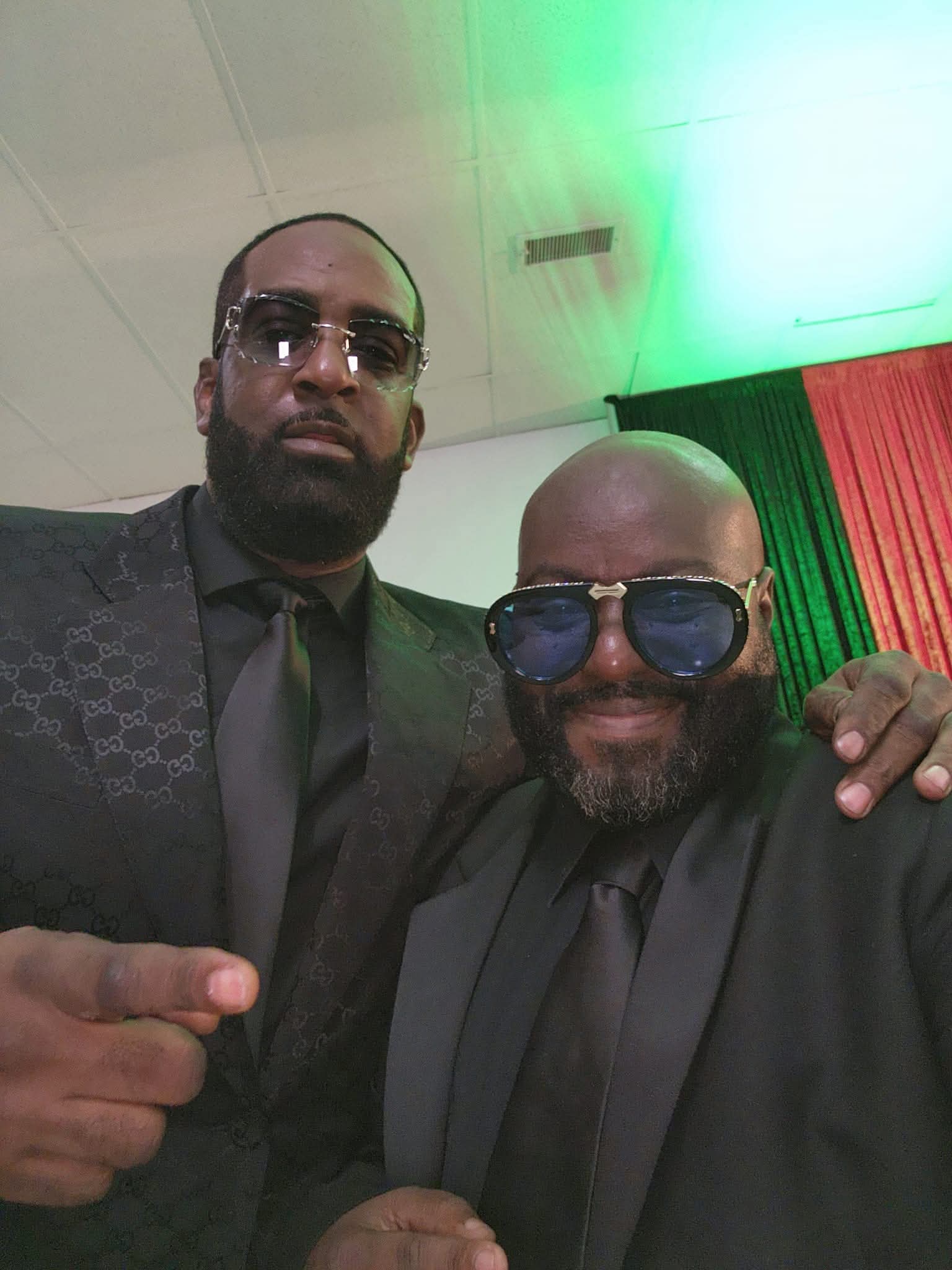 Two men in dark suits and sunglasses pose together indoors with green and red curtains behind them.