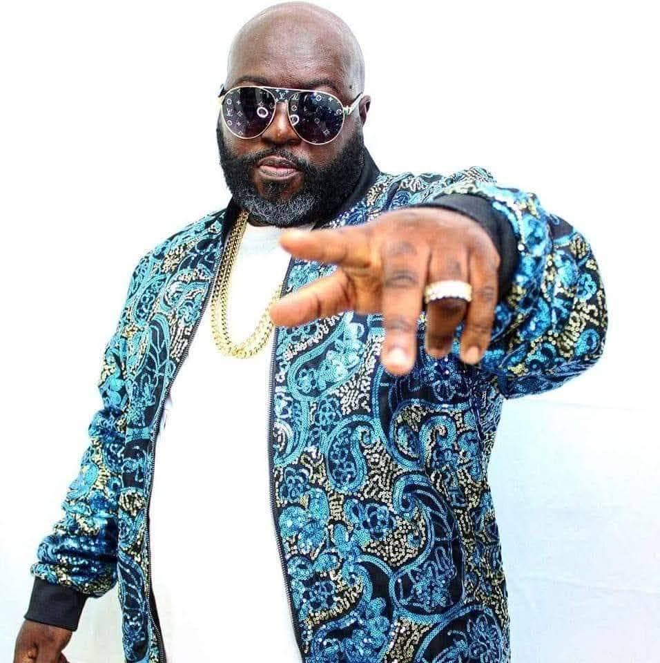 Man with sunglasses and a patterned blue jacket points forward, wearing a gold chain and ring, against a white background.
