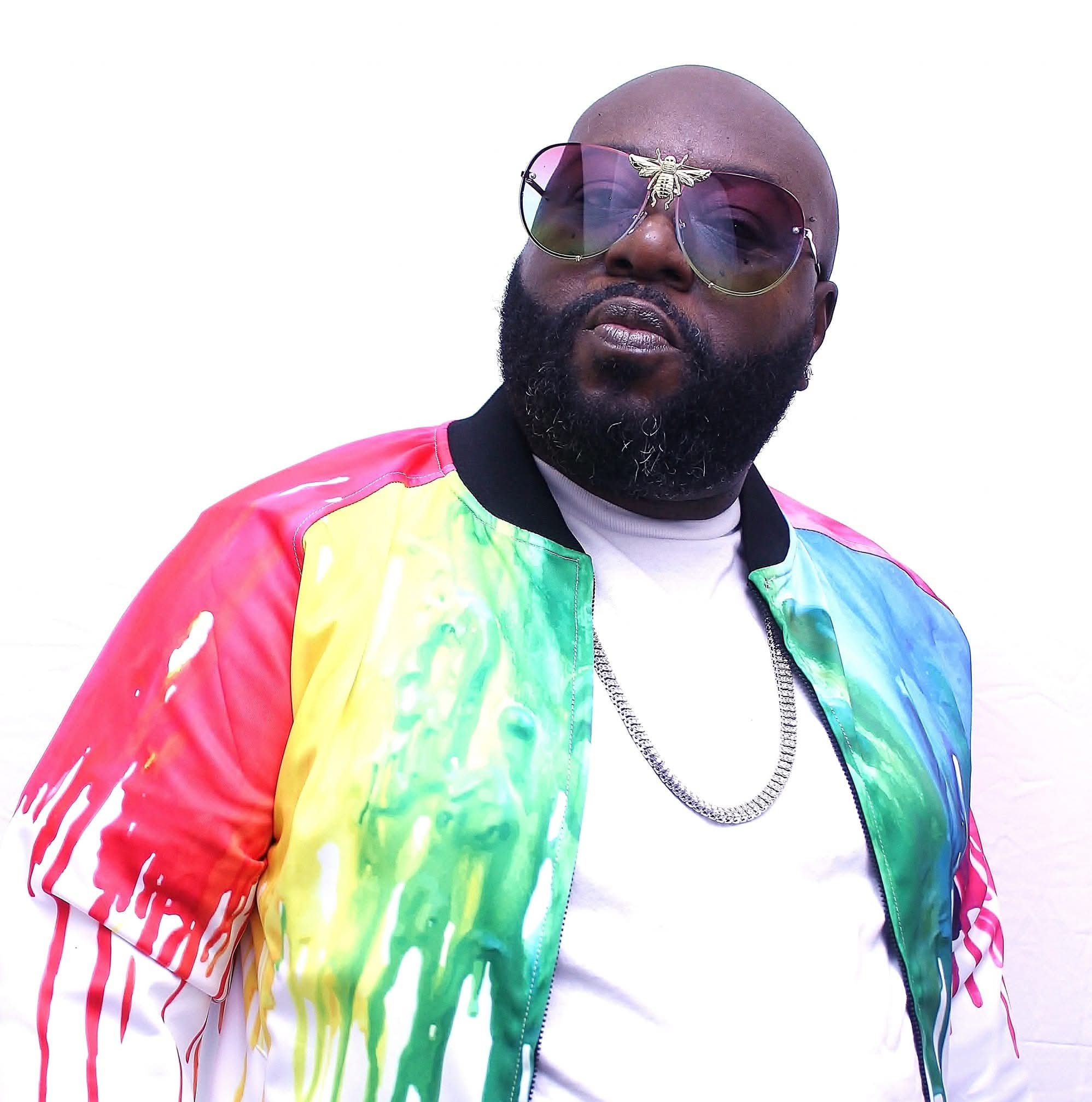 Man with sunglasses, beard, wearing a colorful tie-dye jacket and white shirt, against a white background.