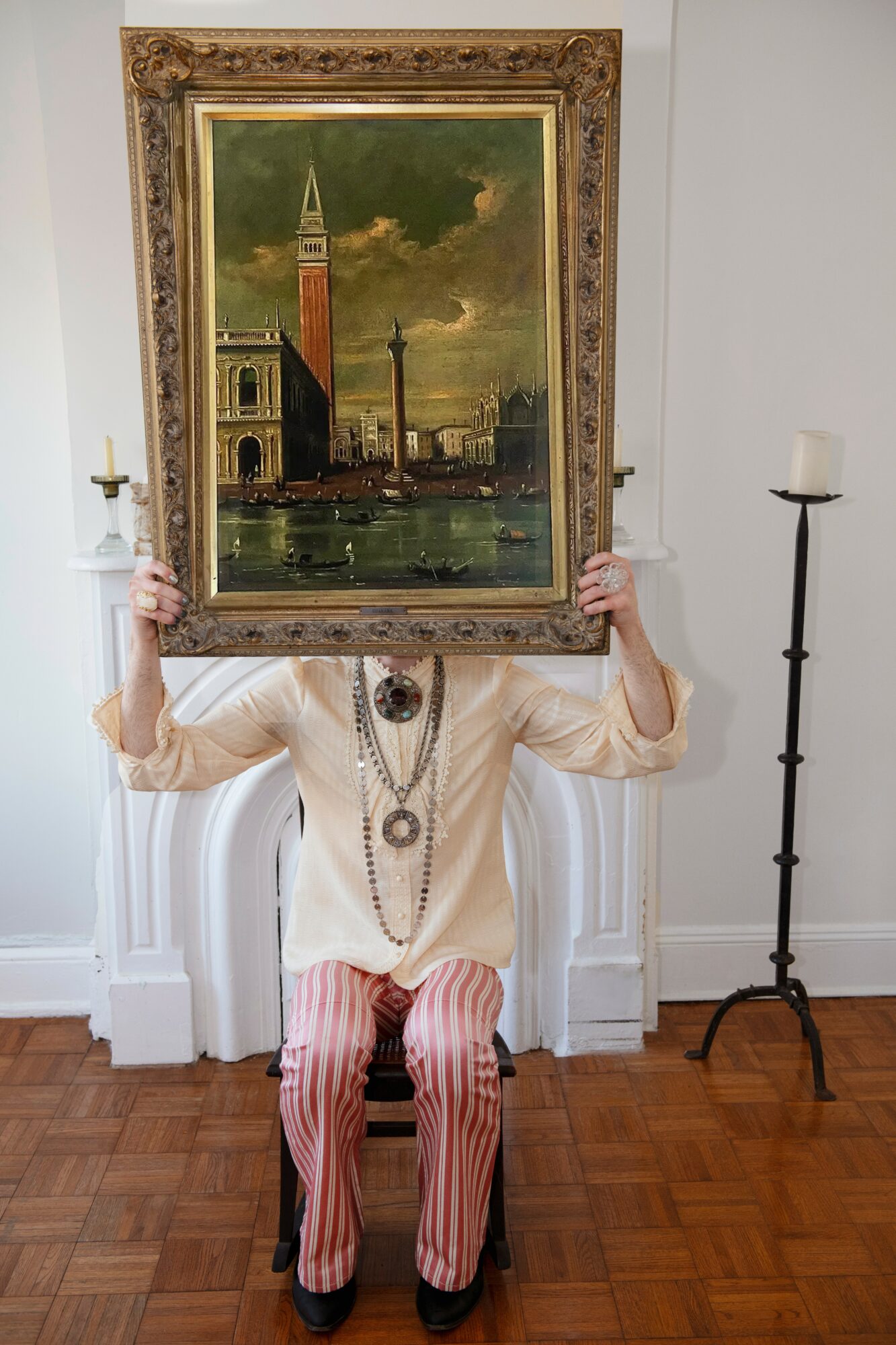Person holding a framed painting of Venice with a canal, buildings, and a cloudy sky, covering their face, sitting on a white bench.