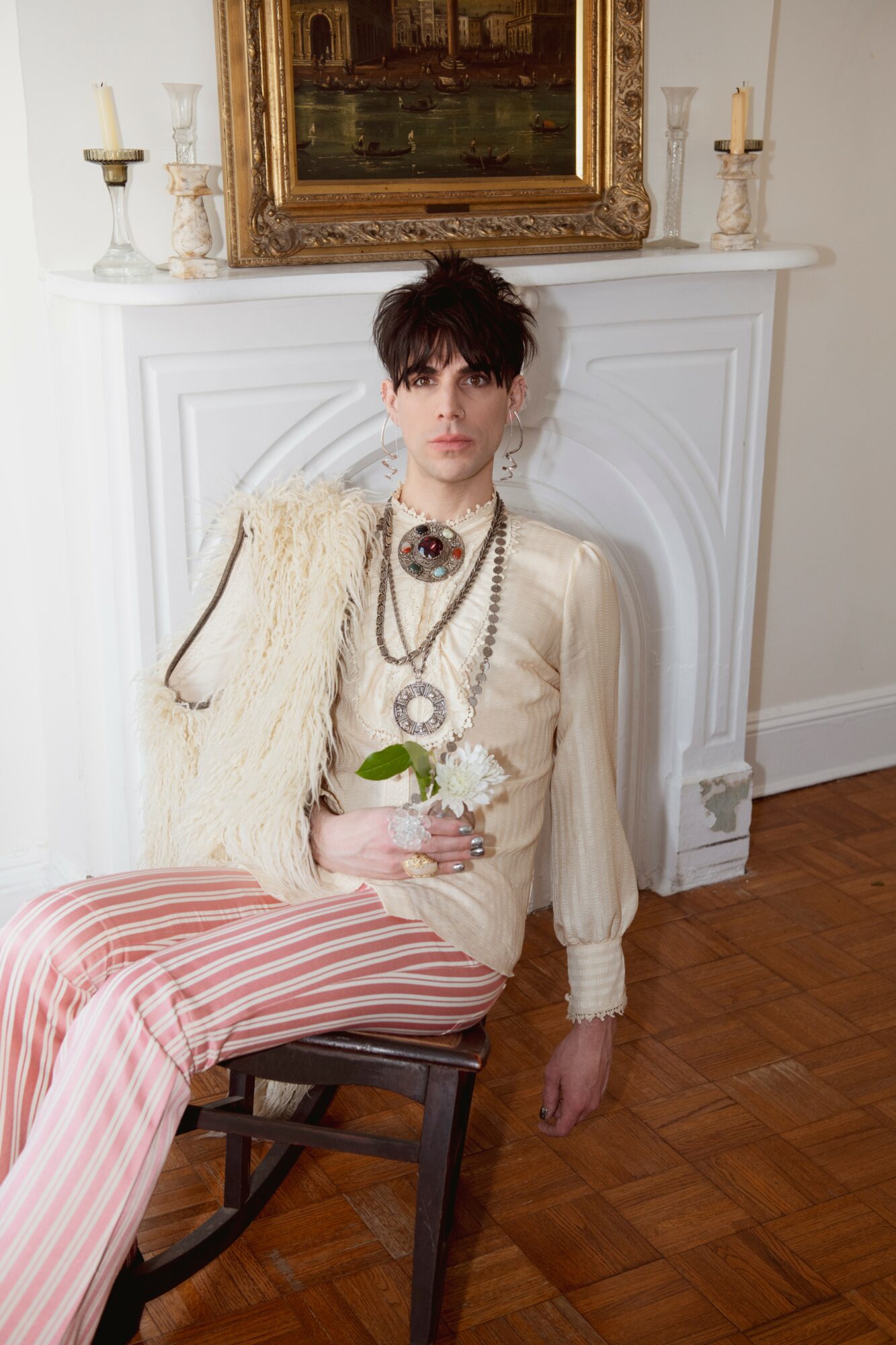 Young woman with dark hair and makeup, sitting on a chair in front of a white fireplace, holding a flower, wearing vintage-style clothing.