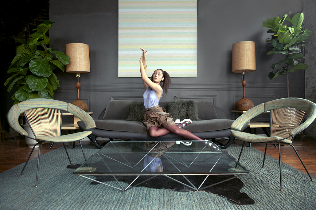 Young woman sitting on a sofa with arms raised, in a modern living room with plants, lamps, and glass coffee table.