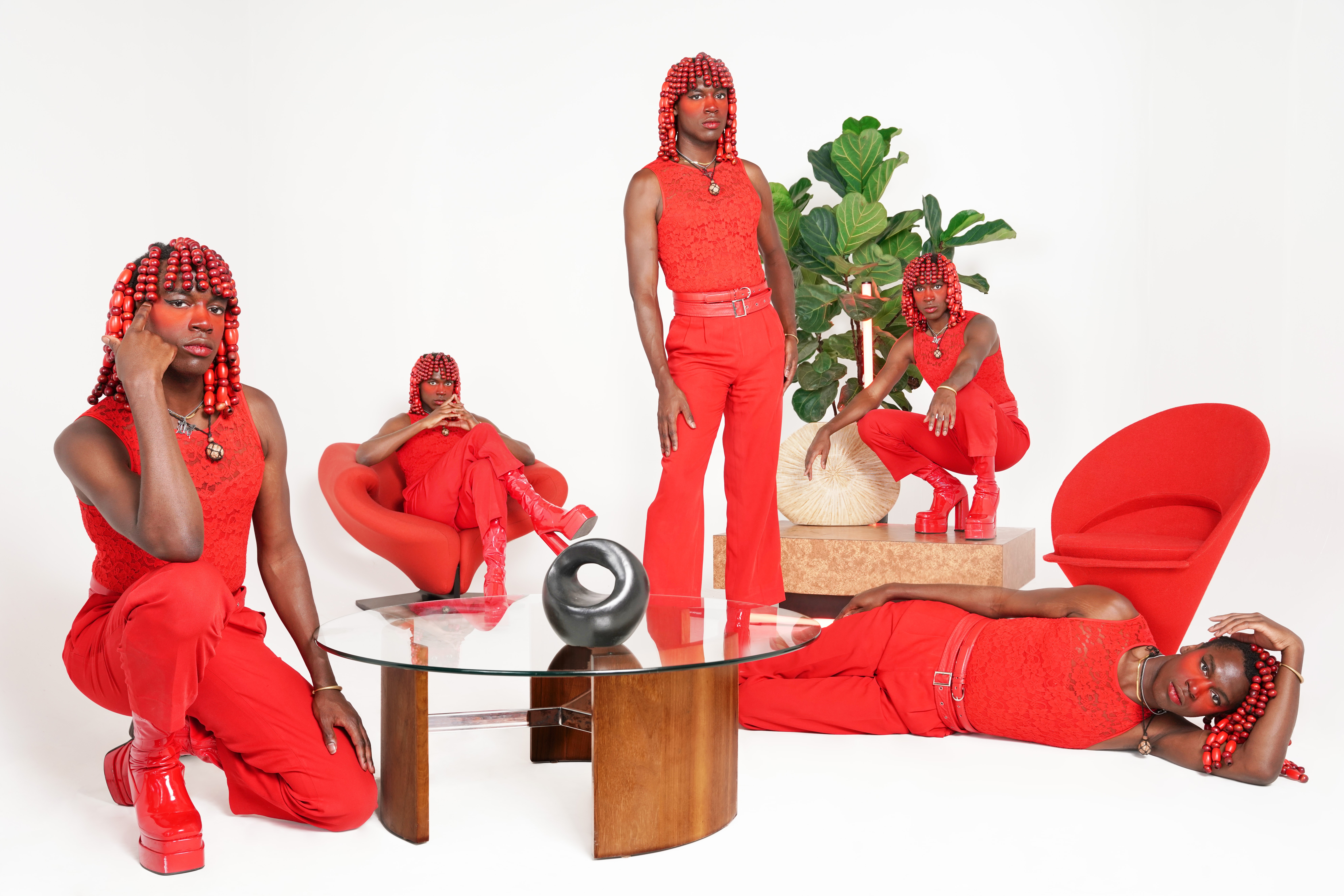 Five women dressed in red, with red hair, in a modern room with a glass table and red chairs, some sitting and one lying down.
