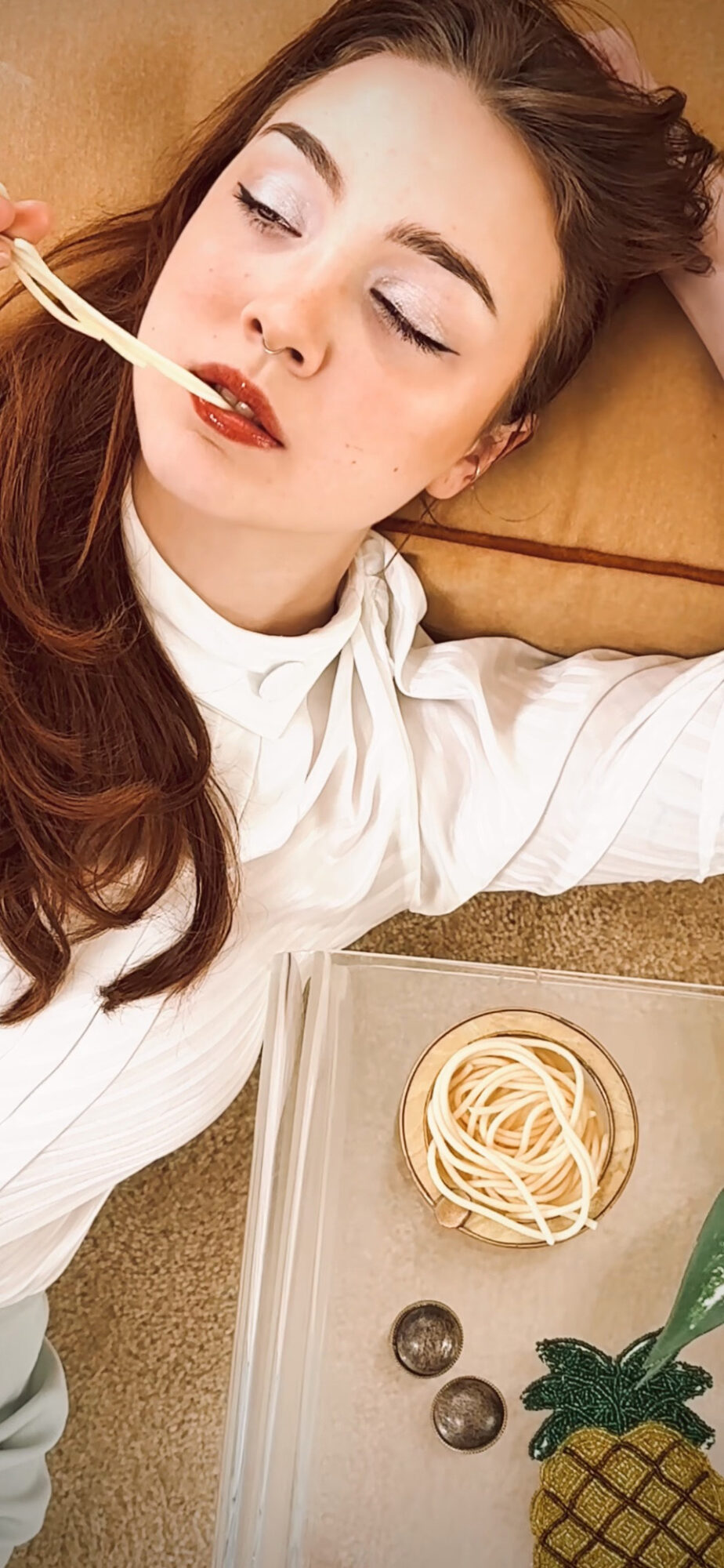 Young woman lying down, eating spaghetti with a fork, with a plate of spaghetti and grapes nearby.